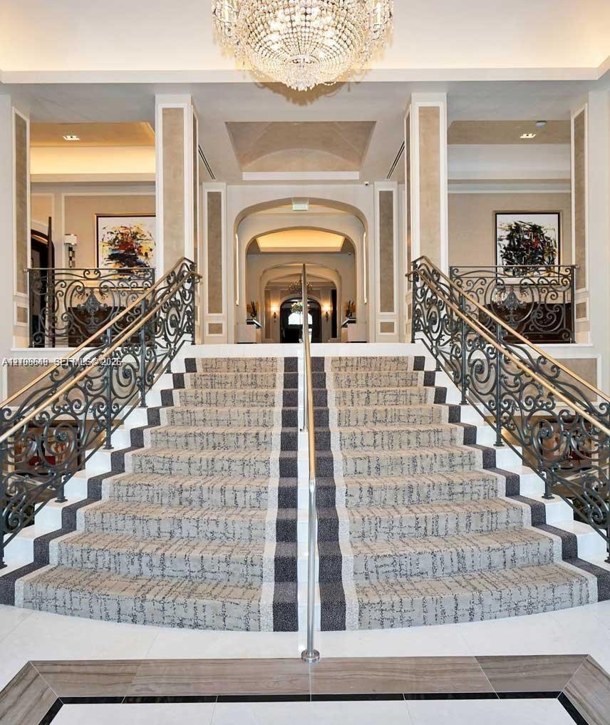 Grand dual staircase featuring intricate wrought iron railings, stone flooring with decorative inlay, and a central crystal chandelier under vaulted c