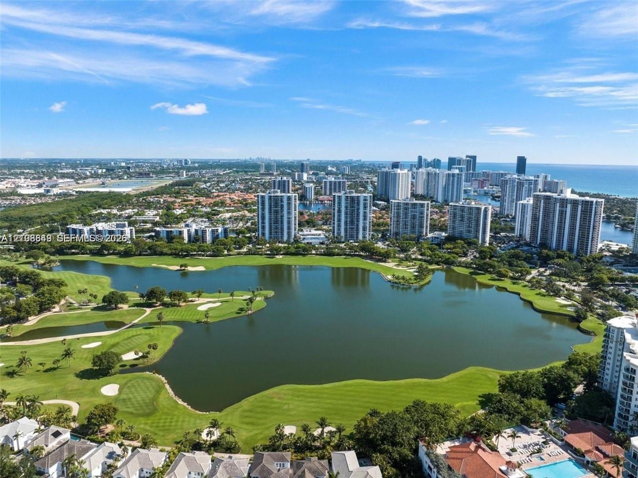 Aerial perspective featuring an expansive lake, manicured golf course greens, sand bunkers, mature landscaping, and coastal high-rise structures