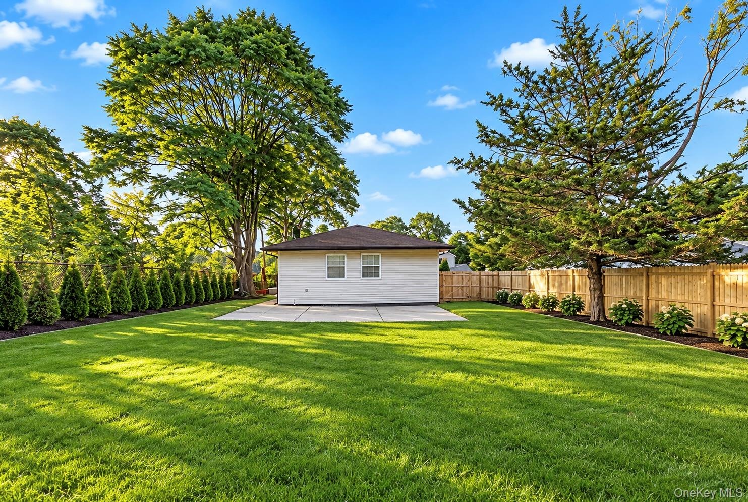 Backyard featuring a manicured lawn, concrete patio, perimeter wood fencing, mature trees, and privacy-screening shrubbery