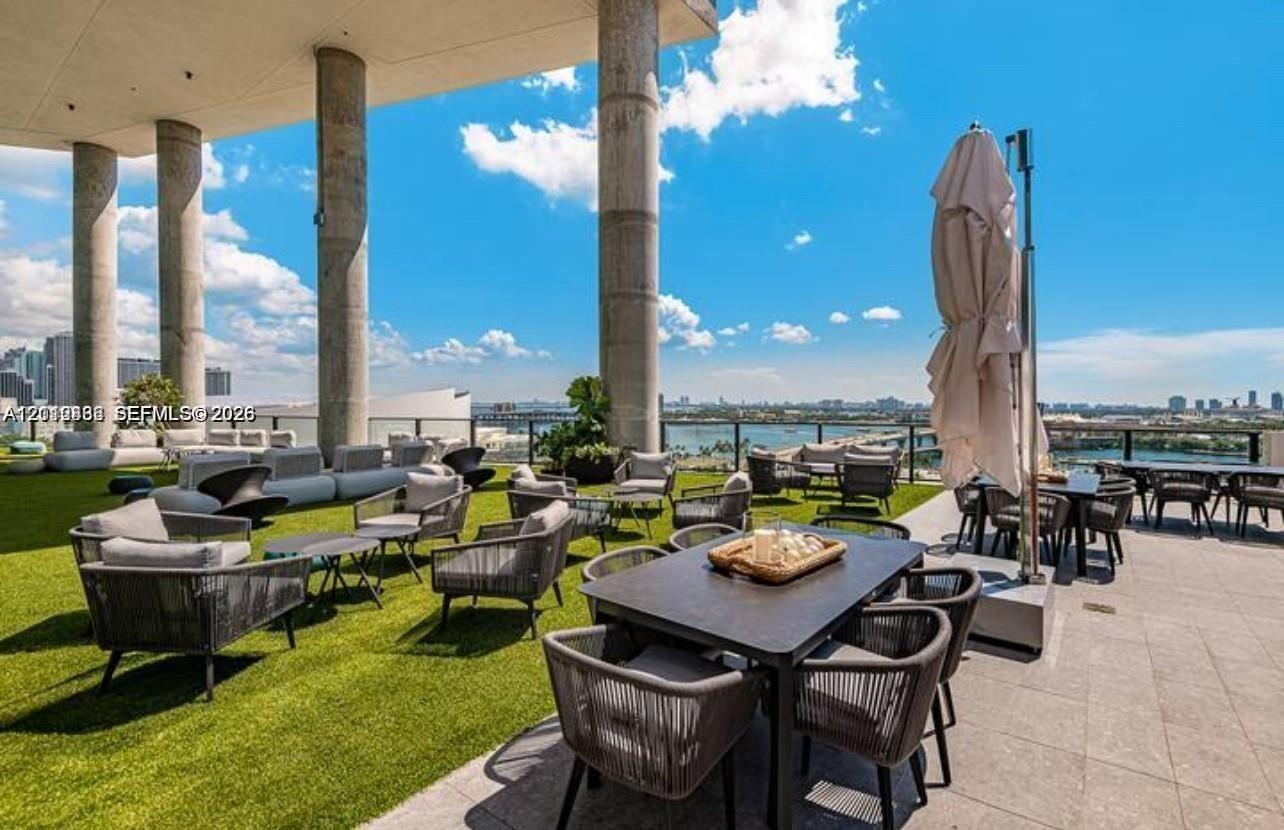 Expansive covered terrace featuring structural concrete columns, synthetic turf surface, stone-tile flooring, and a glass panel perimeter railing