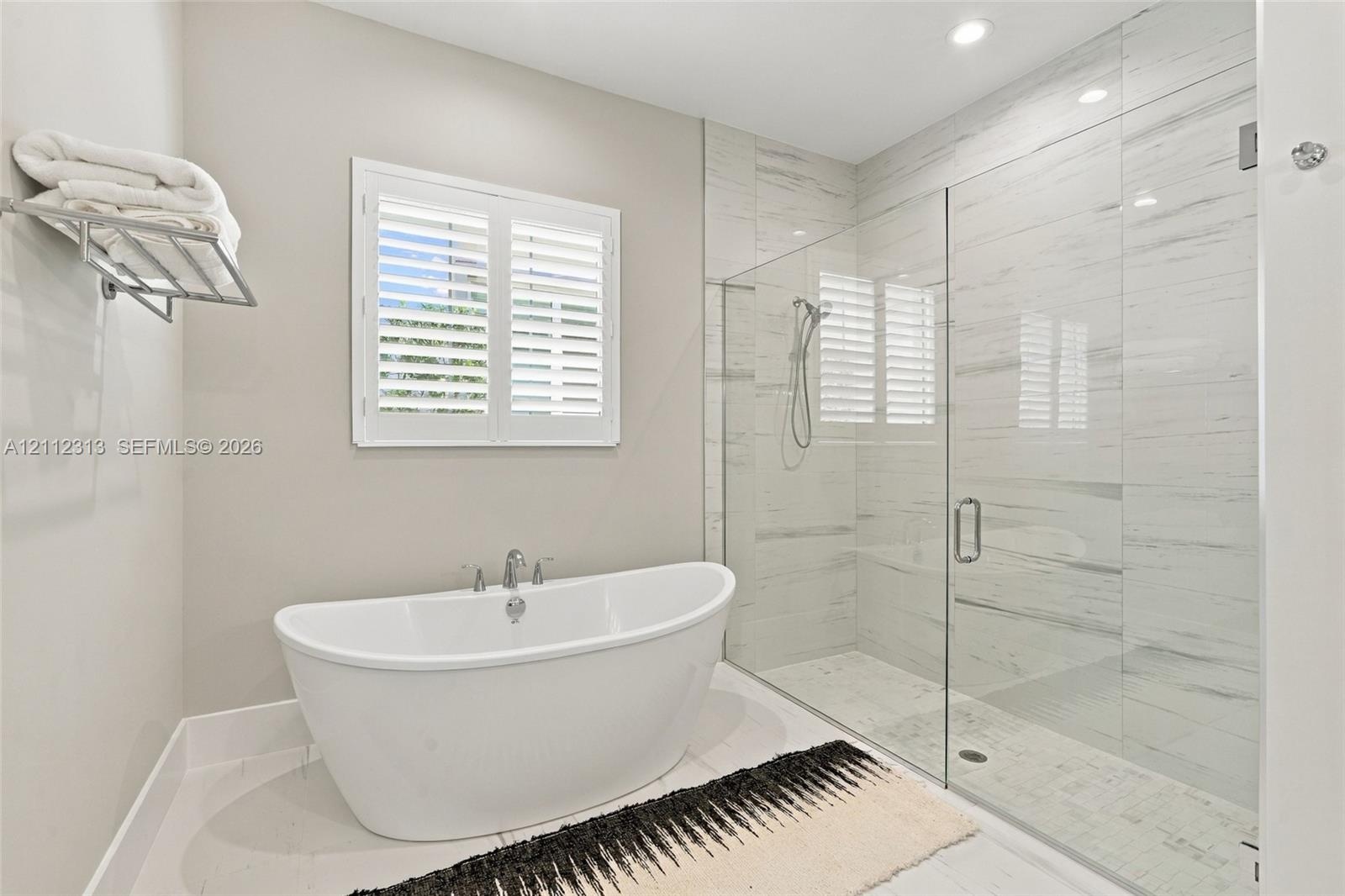 Freestanding soaking tub with chrome fixtures, glass-enclosed walk-in shower with floor-to-ceiling tile, and window shutters