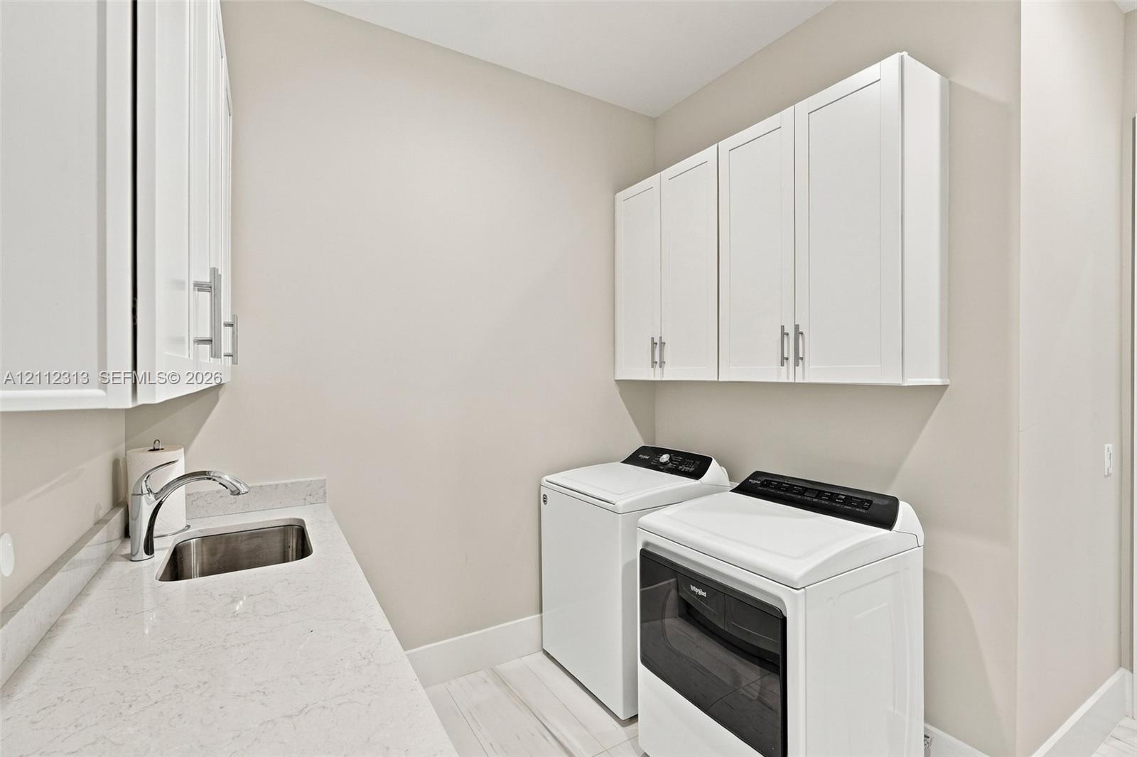 Laundry area featuring white shaker-style wall cabinets, a light-toned countertop, an integrated stainless steel sink with a chrome faucet, and wood-f
