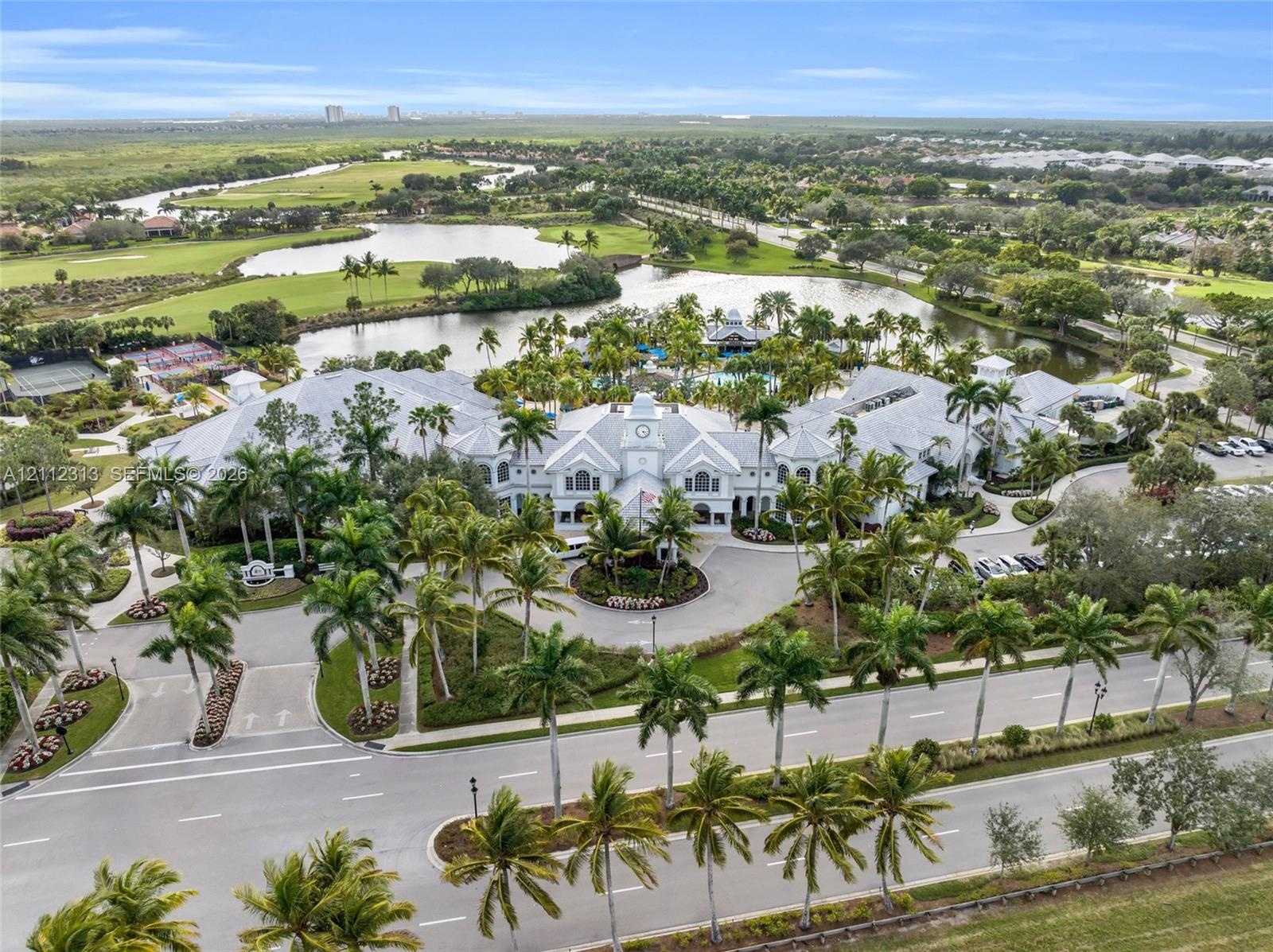 Aerial perspective of a multi-winged facility featuring a central clock tower, expansive pond frontage, mature palm tree landscaping, and paved circul