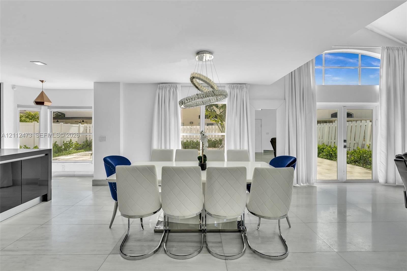 Open-concept interior featuring large-format tile flooring, a contemporary multi-ring crystal chandelier, vaulted ceilings, and dual sets of glass-pan
