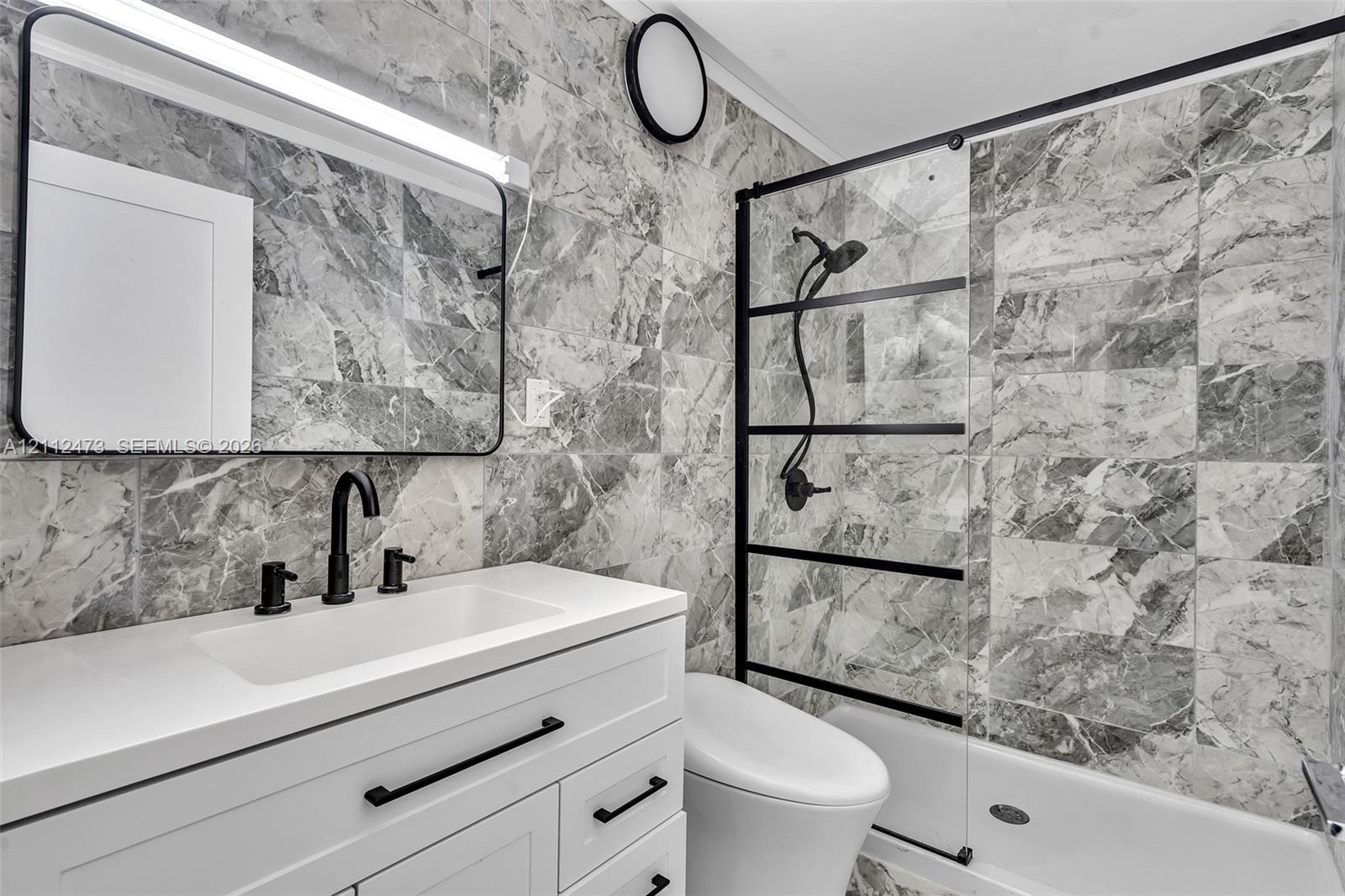 Bathroom featuring full-height stone-finish tile surround, white vanity with quartz countertop, matte black fixtures, framed vanity mirror, and glass 