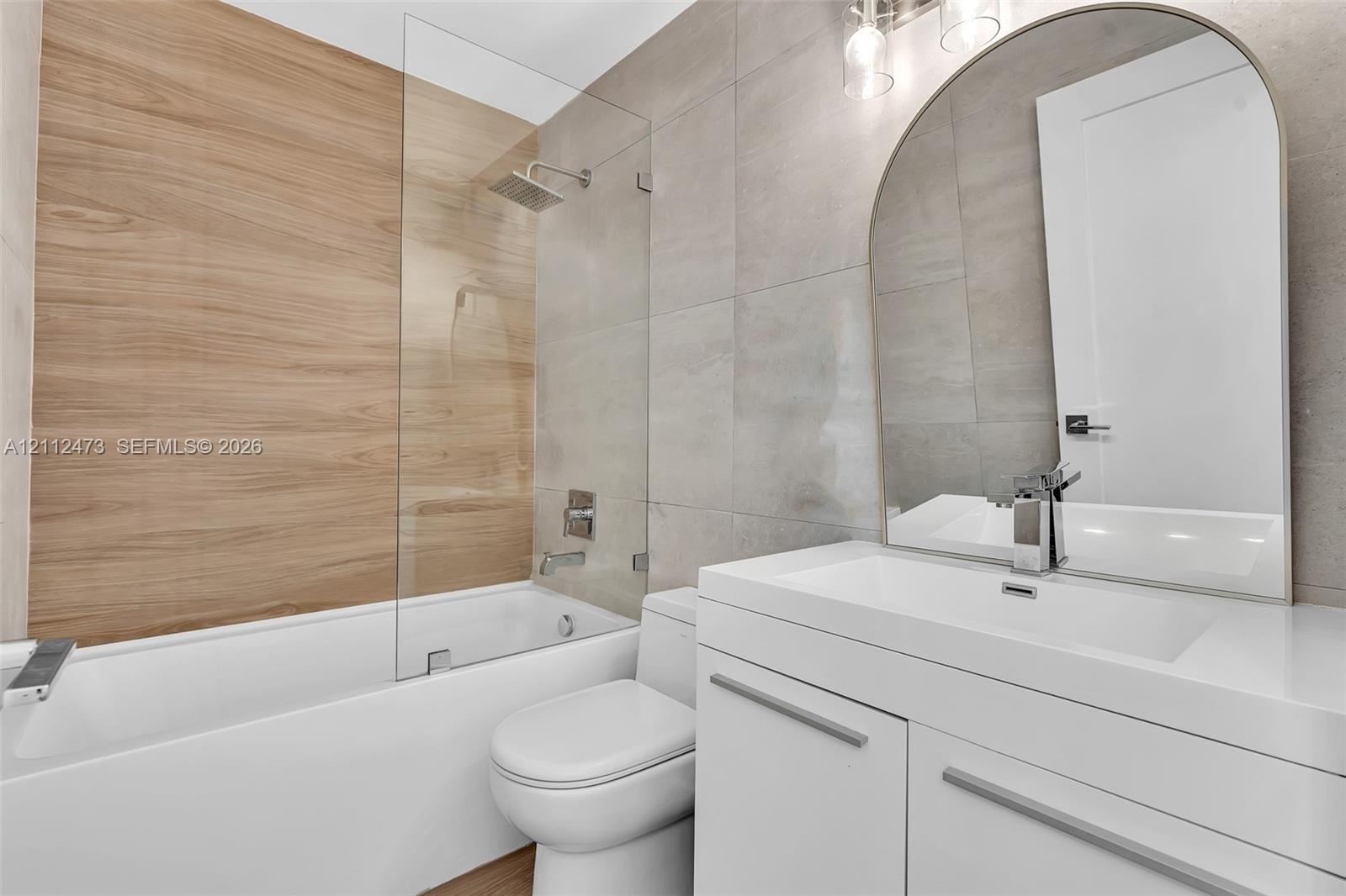 Bathroom featuring a floating white vanity with a rectangular sink, arch-top mirror, wood-finish wall tile, frameless glass shower panel, and large-fo
