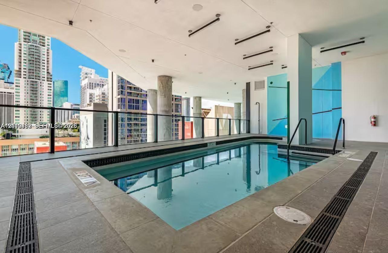 Covered swimming pool featuring large-format tile decking, structural concrete columns, a sleek glass railing, and linear ceiling lighting