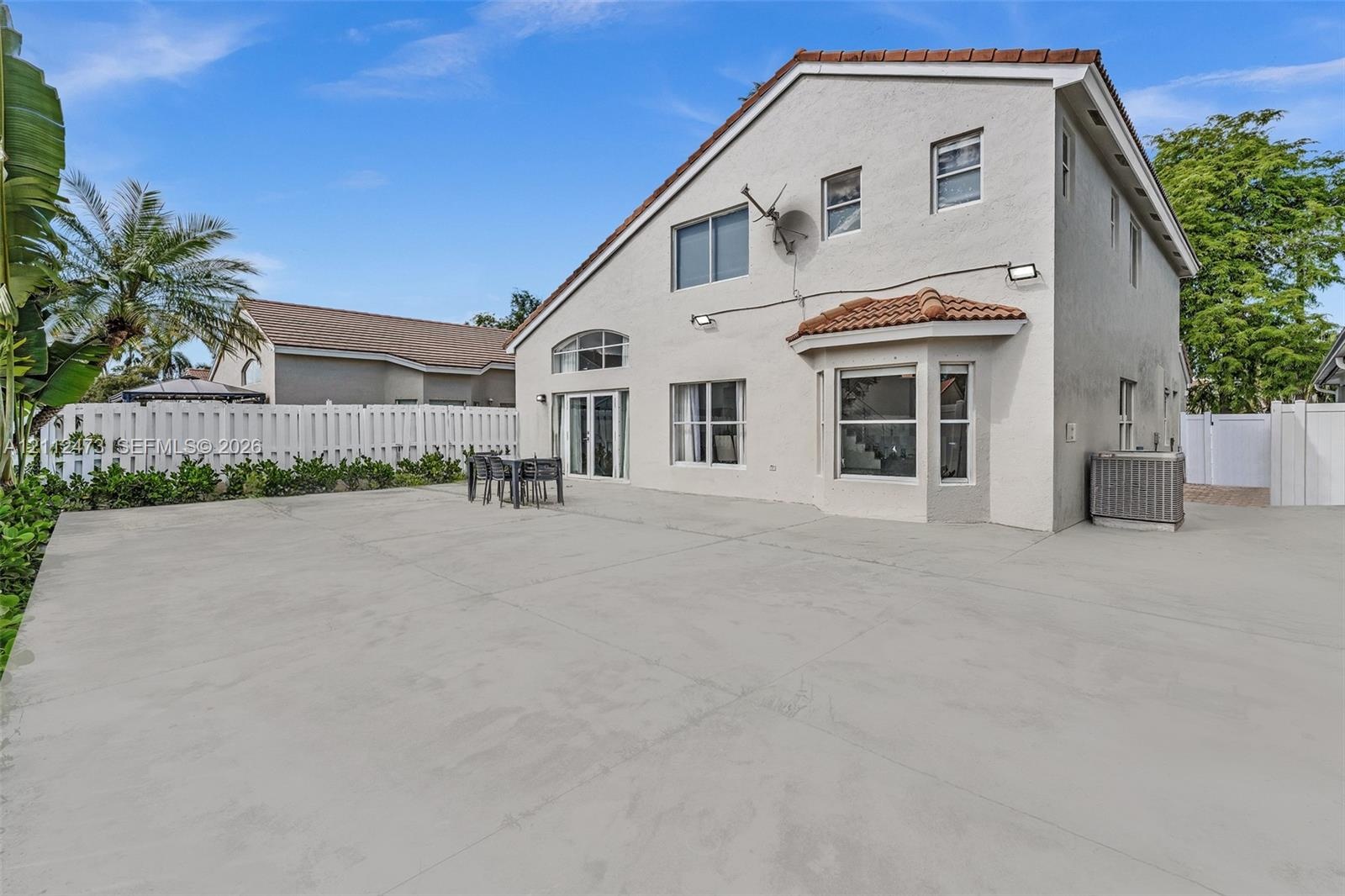 Stucco exterior featuring a tile-accented roof, expansive concrete patio, bay window, and white vinyl privacy fencing