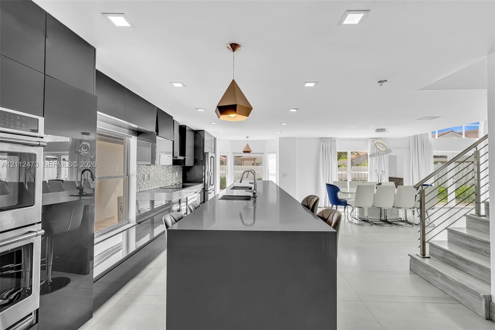 Kitchen featuring high-gloss cabinetry, expansive waterfall-edge island, built-in double ovens, recessed lighting, and large-format tile flooring