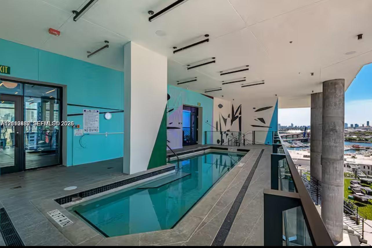 Covered outdoor pool featuring grey stone-finish tile decking, structural concrete support columns, a glass railing system, and linear recessed lighti