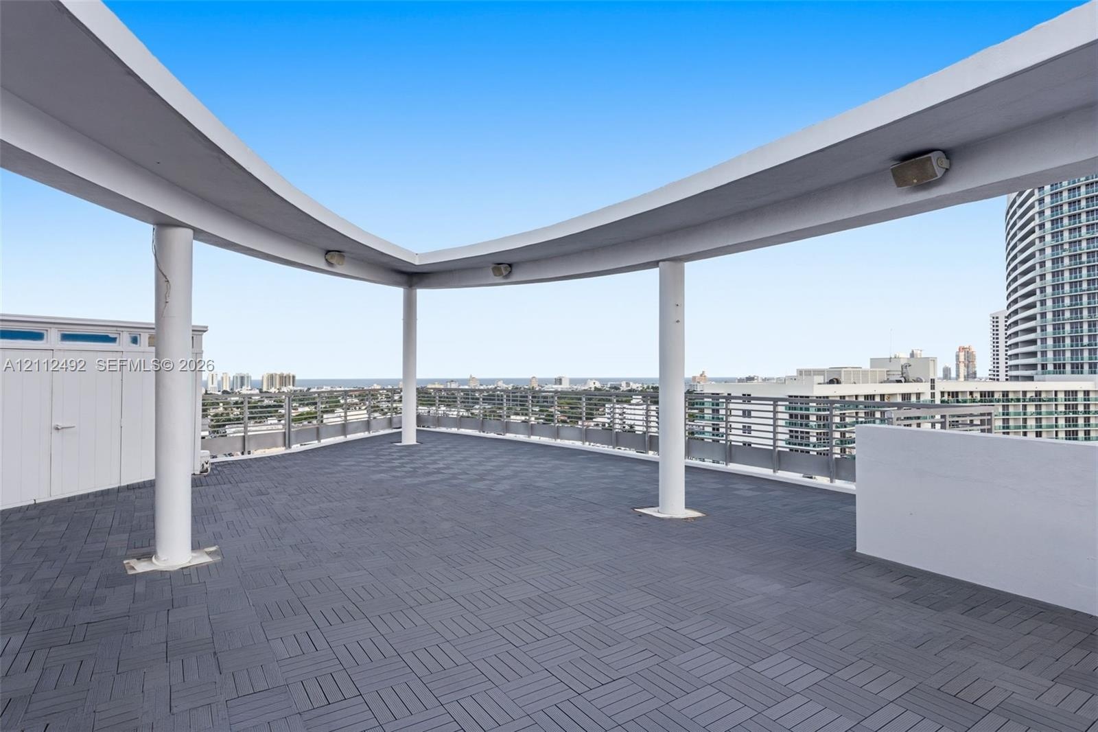 Rooftop terrace featuring dark modular interlocking flooring, a curved architectural canopy supported by cylindrical columns, and metal perimeter rail