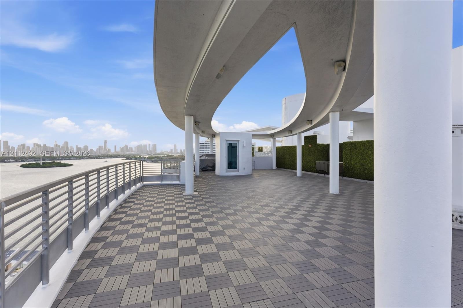 Expansive outdoor terrace featuring a geometric tile deck, curved architectural overhang, cylindrical support columns, metal railing, and privacy hedg