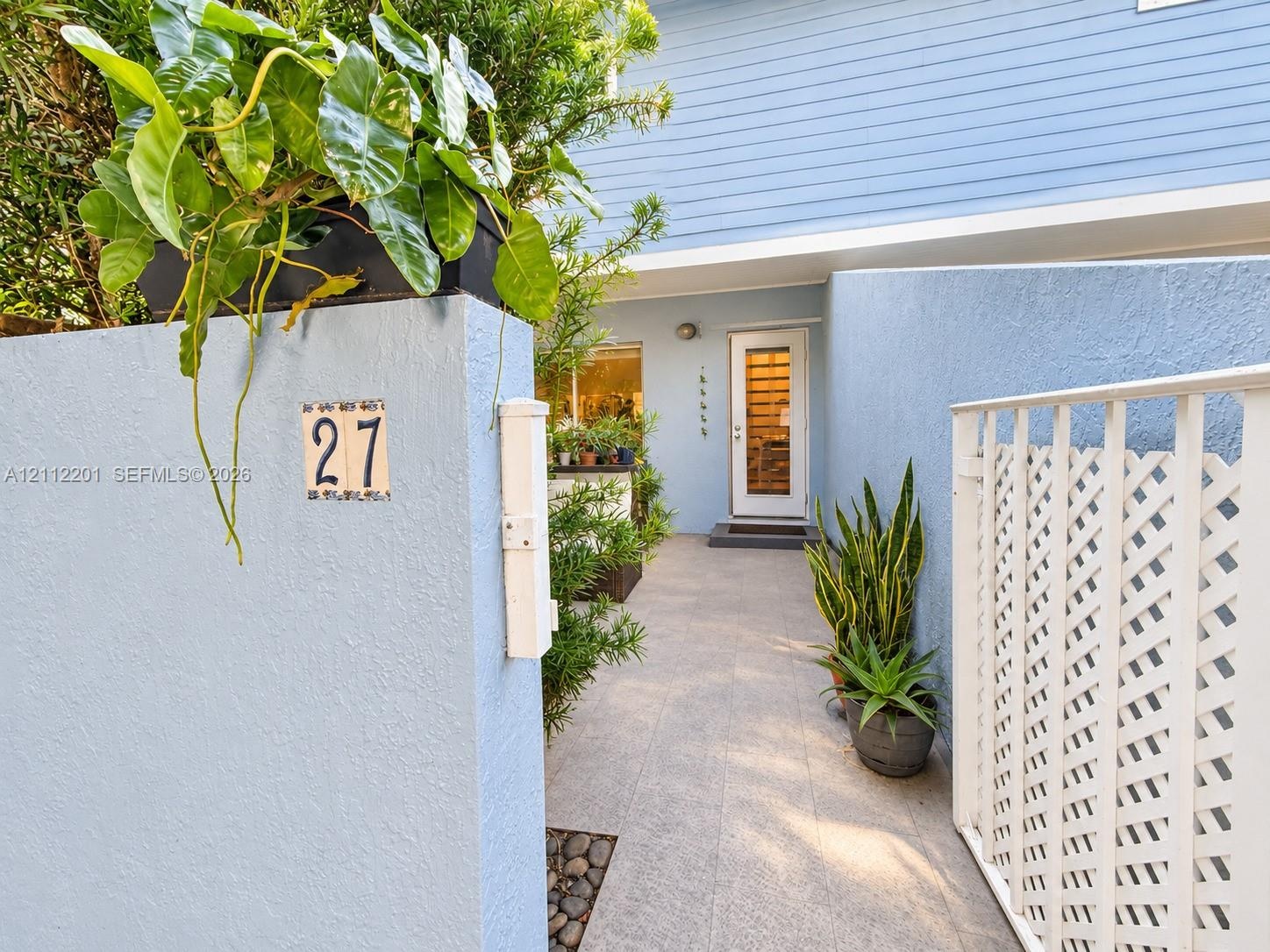 Private entryway featuring stucco perimeter walls, horizontal lap siding, tiled walkway, and a white lattice fence