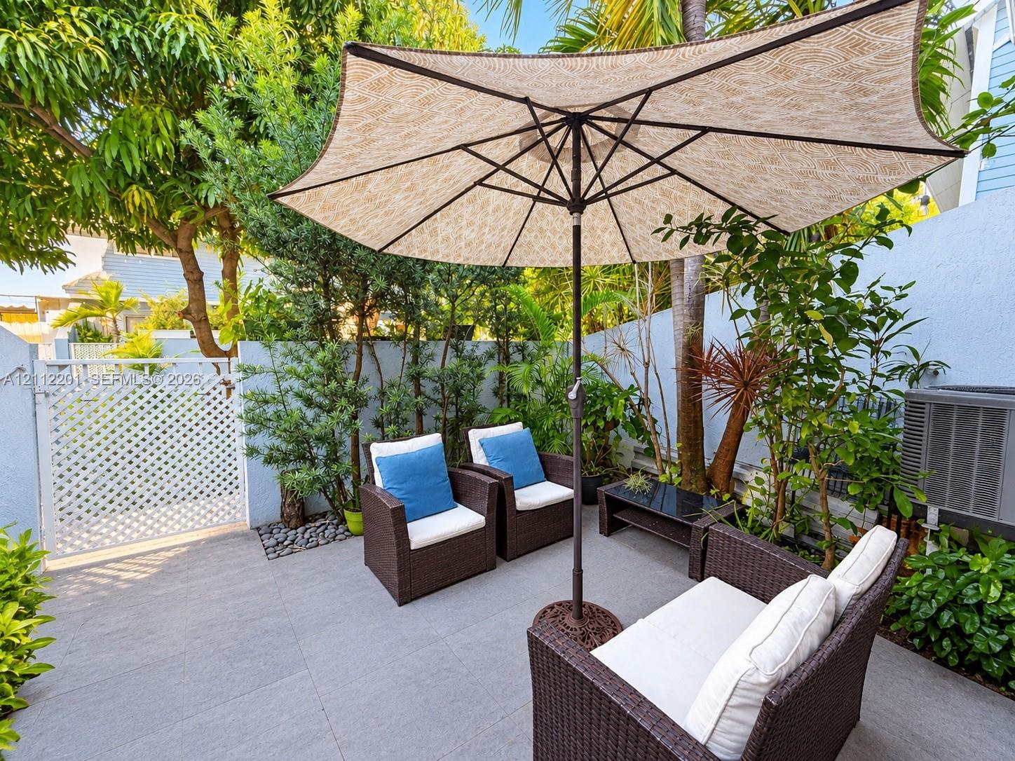 Private patio featuring grey tile flooring, solid perimeter walls, and a decorative white lattice gate surrounded by established landscaping