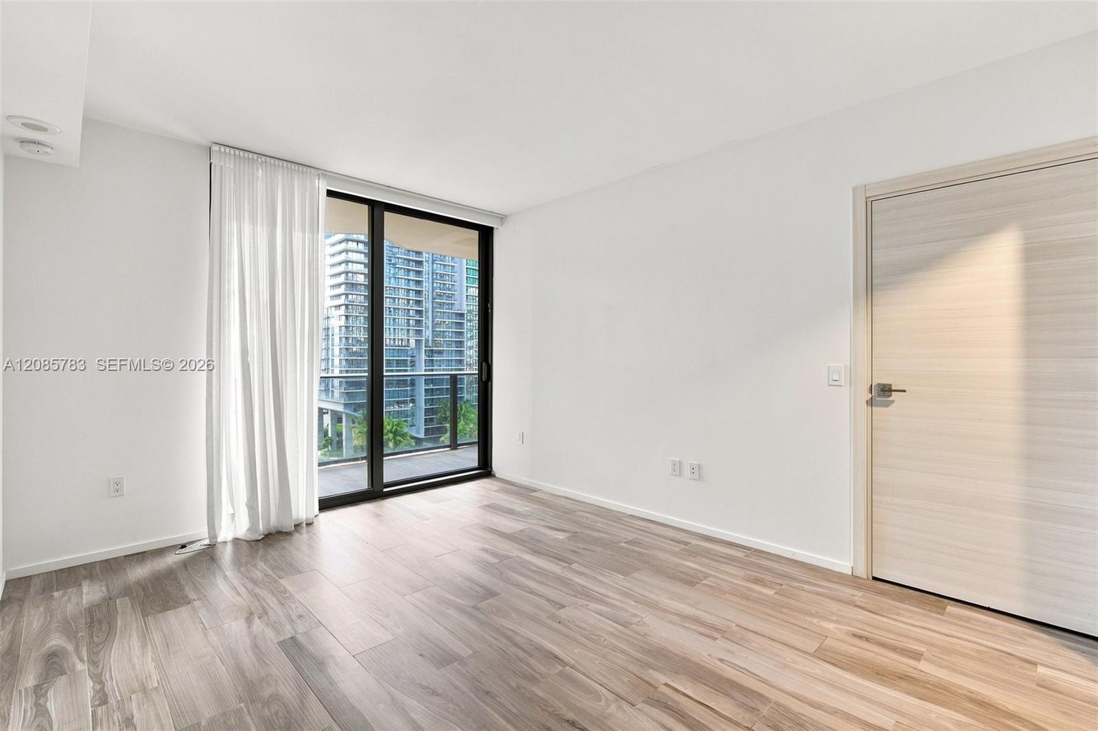 Room featuring wood-finish flooring, floor-to-ceiling glass sliding door, balcony access, and a light-toned wood-finish interior door