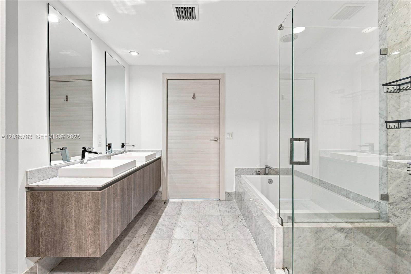 Floating wood-finish vanity with dual vessel sinks, stone-finish countertops, frameless glass shower enclosure, deep soaking tub, and stone-finish flo