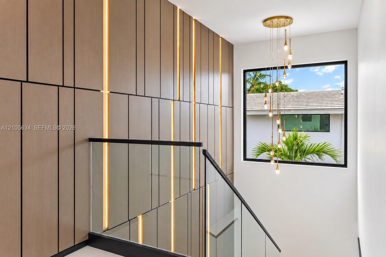 Custom wood-panelled accent wall with integrated vertical lighting, glass railing system featuring a black metal handrail, and multi-pendant brass cha