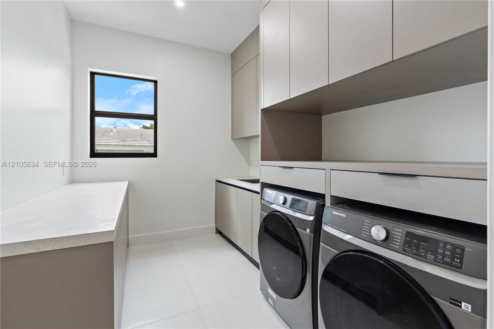 Laundry area featuring custom flat-panel cabinetry, light-toned stone-finish countertops, tile flooring, and integrated laundry appliances