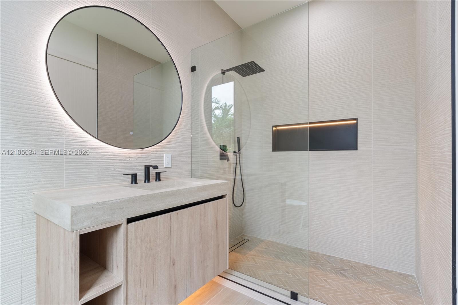 Floating vanity with integrated sink, textured wall tile, circular backlit mirror, frameless glass shower enclosure with rainfall showerhead, and herr