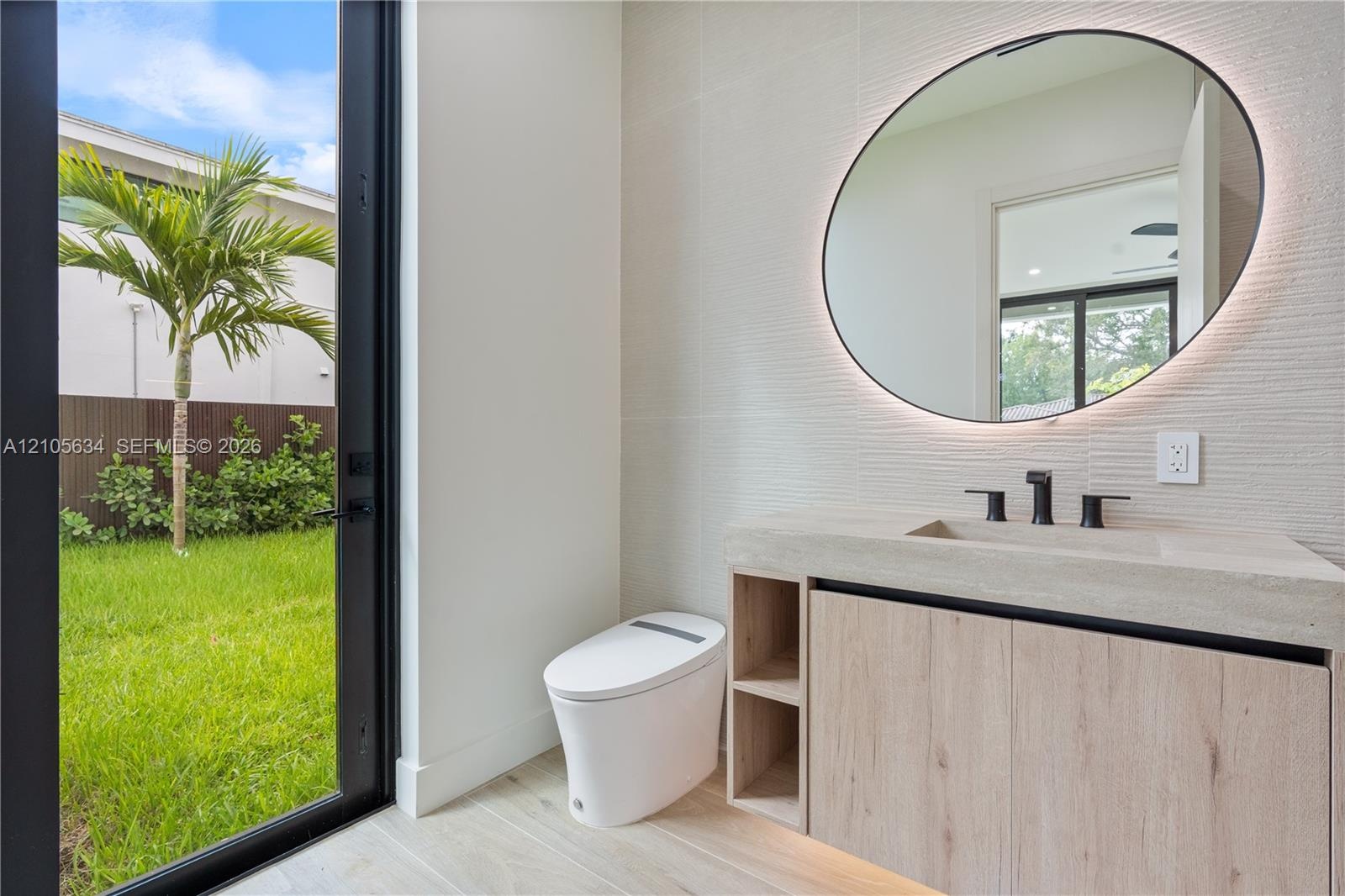 Modern vanity with stone-finish countertop, integrated sink, and wood-finish cabinetry, featuring a circular backlit mirror, wall-mounted matte black 