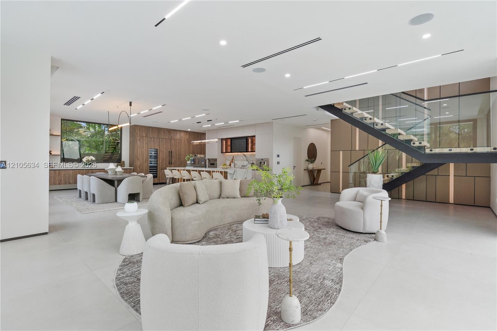 Open-concept floor plan featuring expansive stone-tile flooring, recessed linear lighting, custom wood-paneled cabinetry, and a floating staircase wit