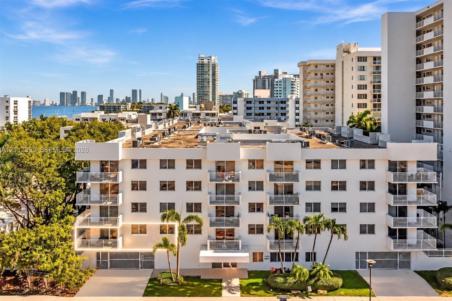 Mid-rise residential building featuring white stucco facade, multiple private balconies with railings, and mature palm tree landscaping