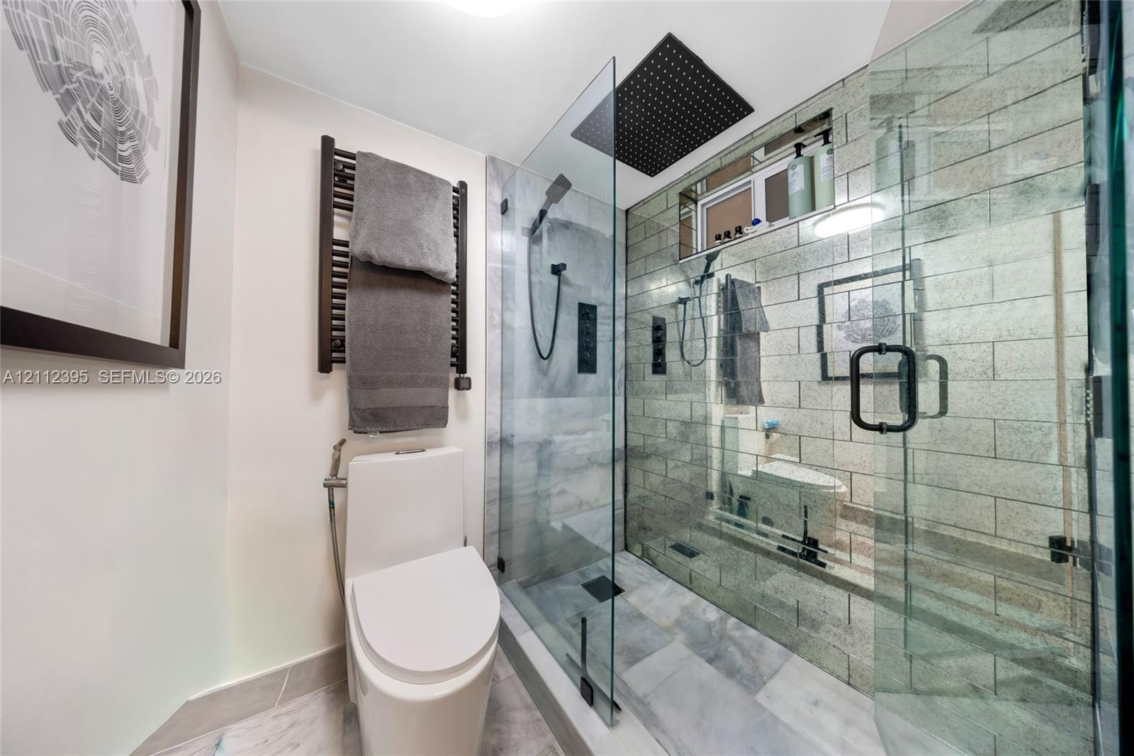 Walk-in shower with stone-tile surround, glass enclosure, and rainfall showerhead, complemented by a matte black heated towel rail and wall-mounted pl