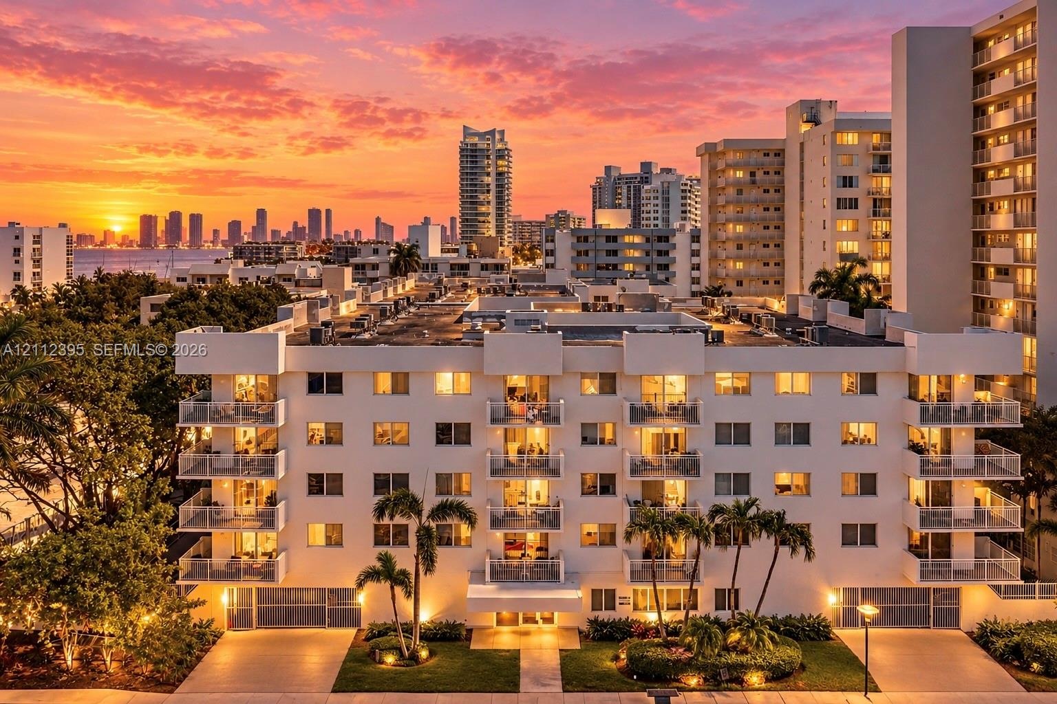 Mid-rise residential building featuring stacked private balconies with glass railings, landscaped grounds with mature palm trees, and exterior hardsca