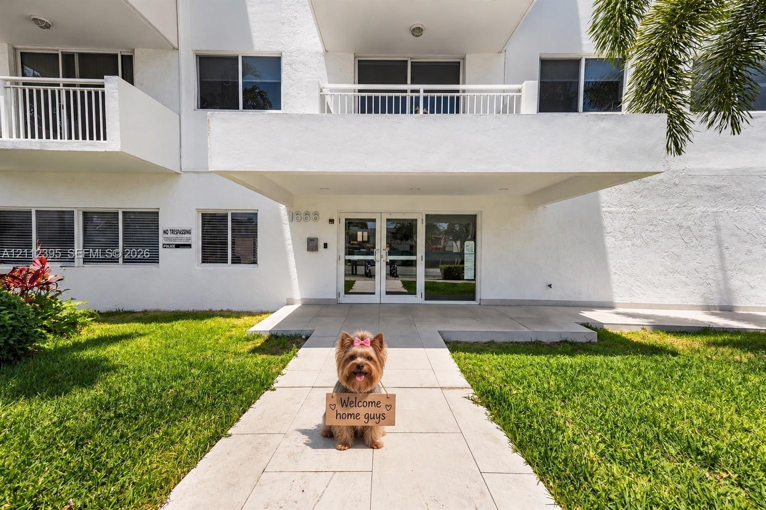 Multi-story residential building exterior featuring a covered entryway, dual glass-panel entrance doors, second-level balconies with white railings, a
