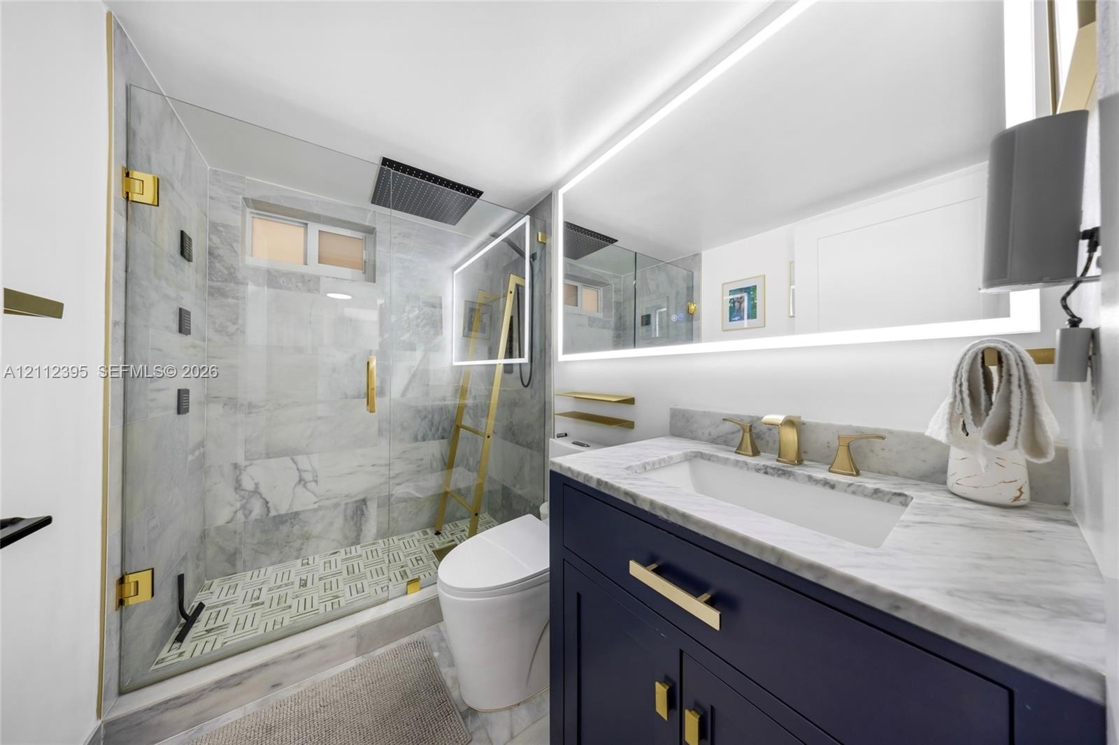 Modern vanity with navy cabinetry, stone countertop, and brass hardware, featuring a glass-enclosed shower with floor-to-ceiling stone tiling, overhea