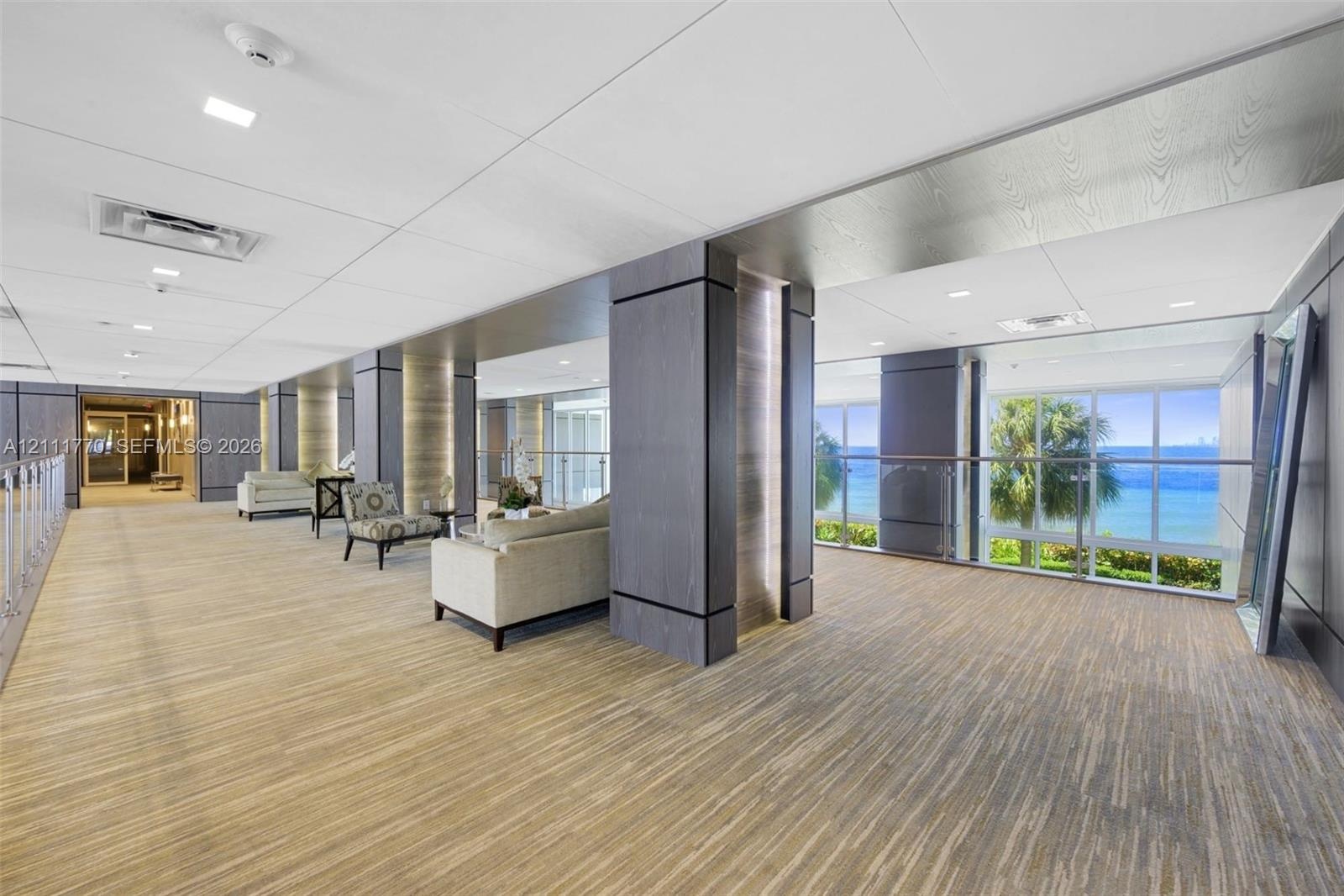 Expansive common area featuring wood-finish flooring, floor-to-ceiling windows with ocean scenery, dark wood-tone architectural columns, and recessed 