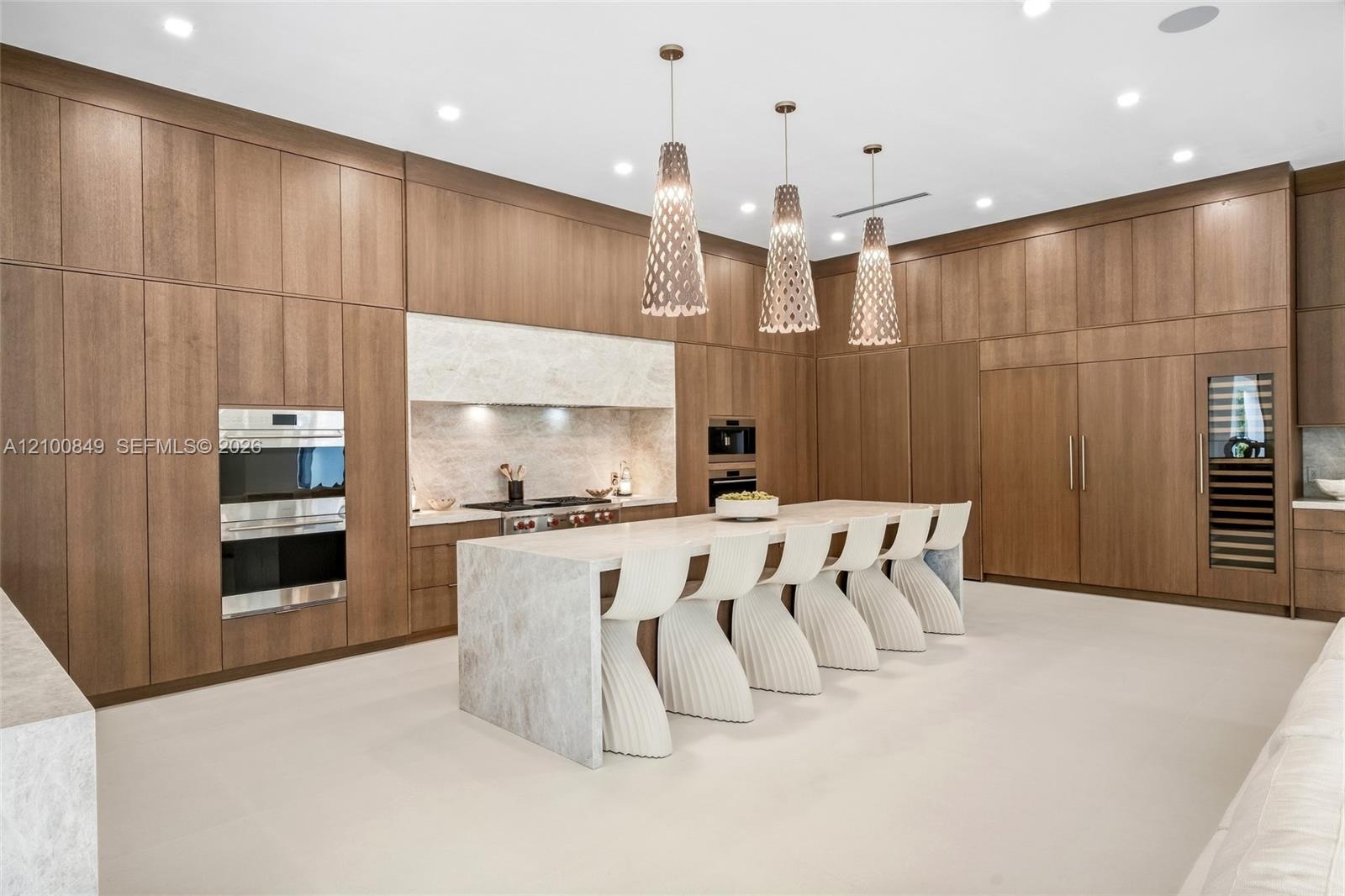 Floor-to-ceiling cabinetry, stone-clad waterfall island, built-in stainless steel double oven, integrated refrigeration, and three sculptural pendant 
