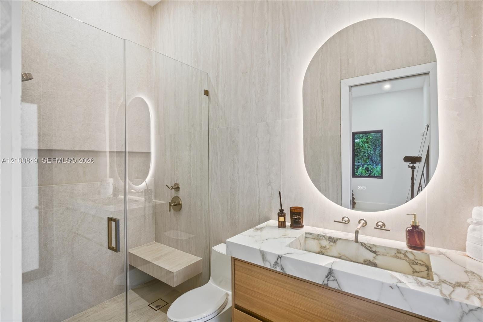 Bathroom featuring a floating wood-finish vanity with a stone countertop, integrated sink, illuminated arched mirror, and a walk-in shower with floor-