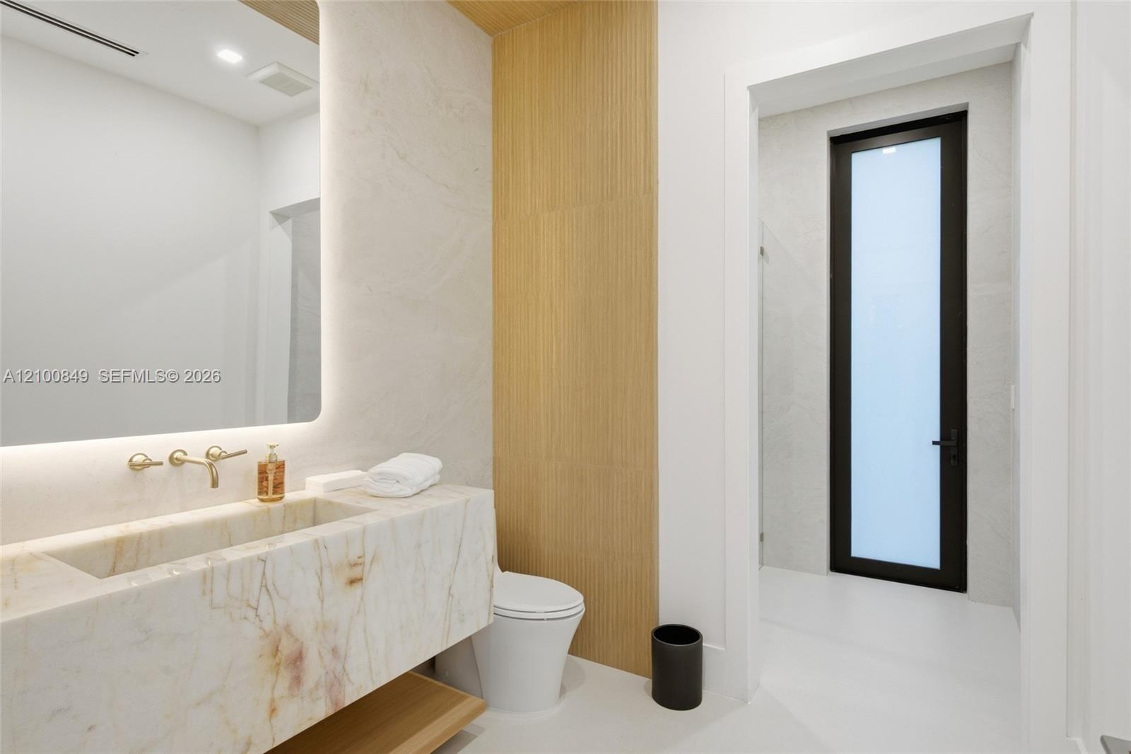Floating stone vanity with integrated basin, wall-mounted brass faucet, illuminated recessed mirror, and wood-panel accent wall