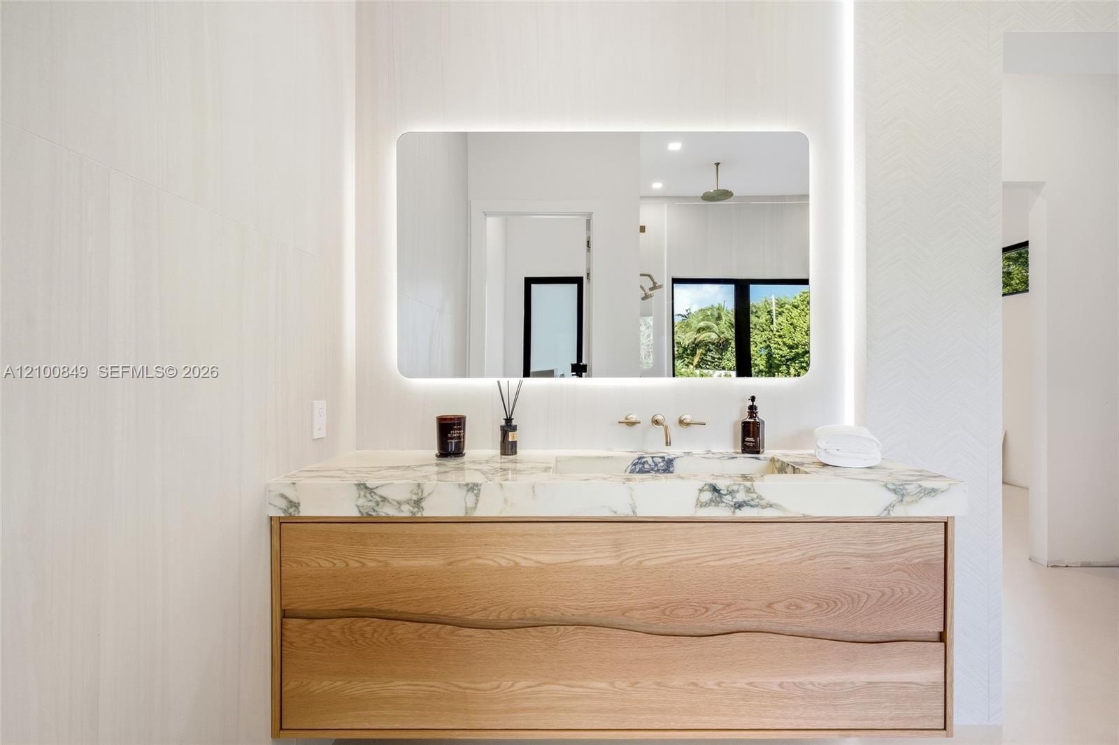 Floating vanity with natural-finish wood cabinetry, stone-top counter with integrated sink, wall-mounted metallic faucet, and illuminated rectangular 