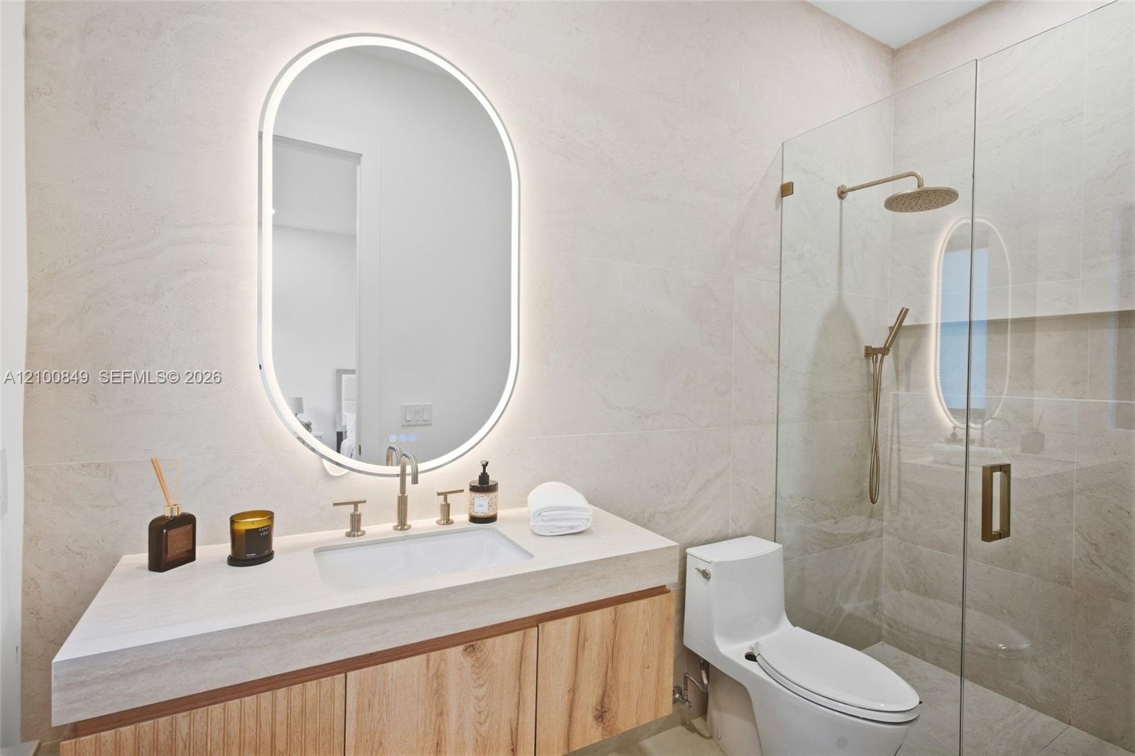 Floating wood-finish vanity with an integrated basin, illuminated oval mirror, stone-tiled walls, and a glass-enclosed shower with a rainfall showerhe