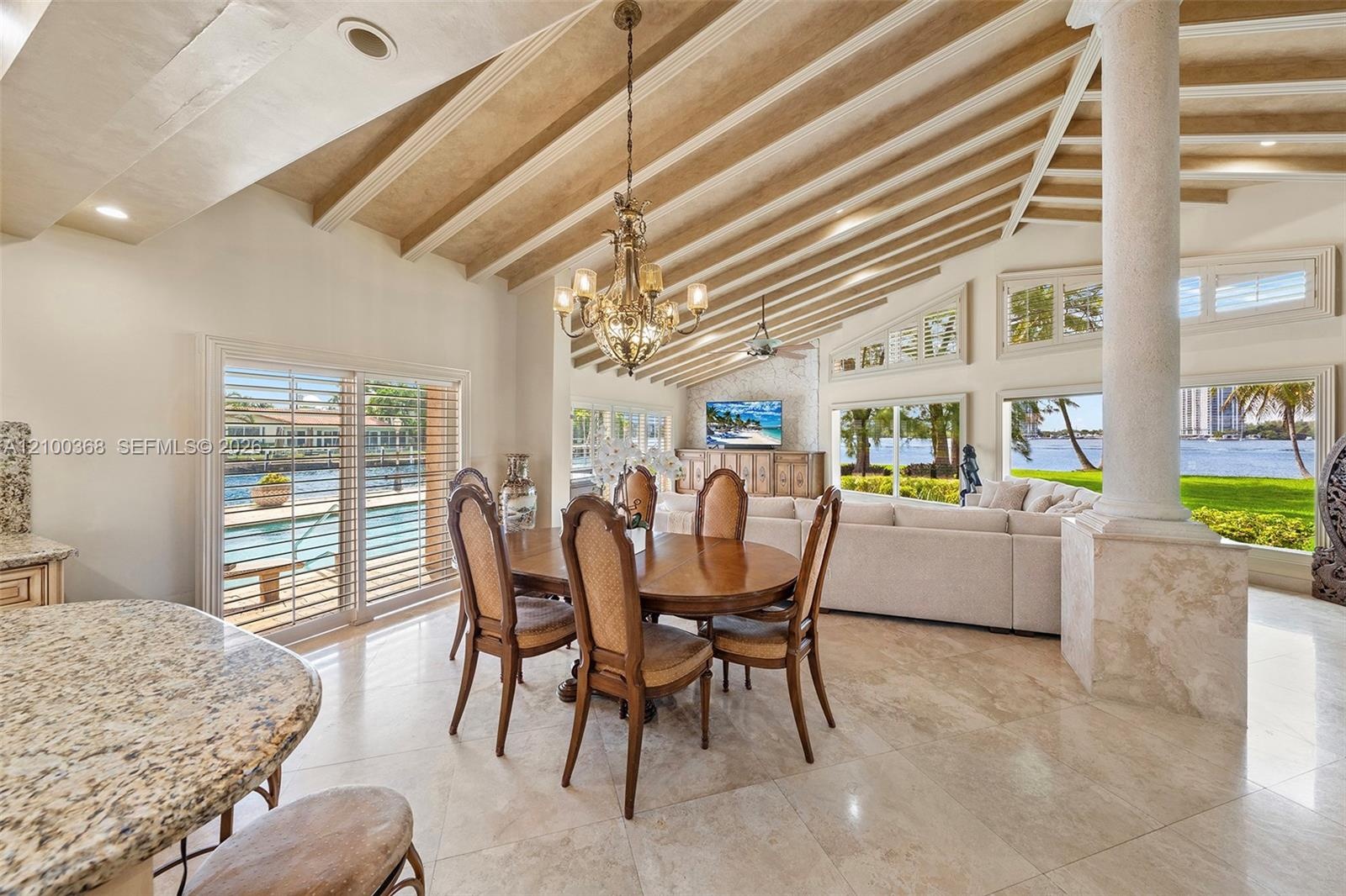 Open-concept living space featuring vaulted ceilings with exposed wood beams, natural stone flooring, a decorative stone column, and expansive floor-t