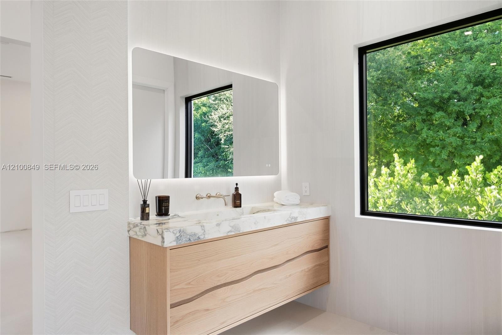 Floating wood-finish vanity with a stone countertop, wall-mounted faucet, integrated LED-backlit mirror, and large window with exterior greenery