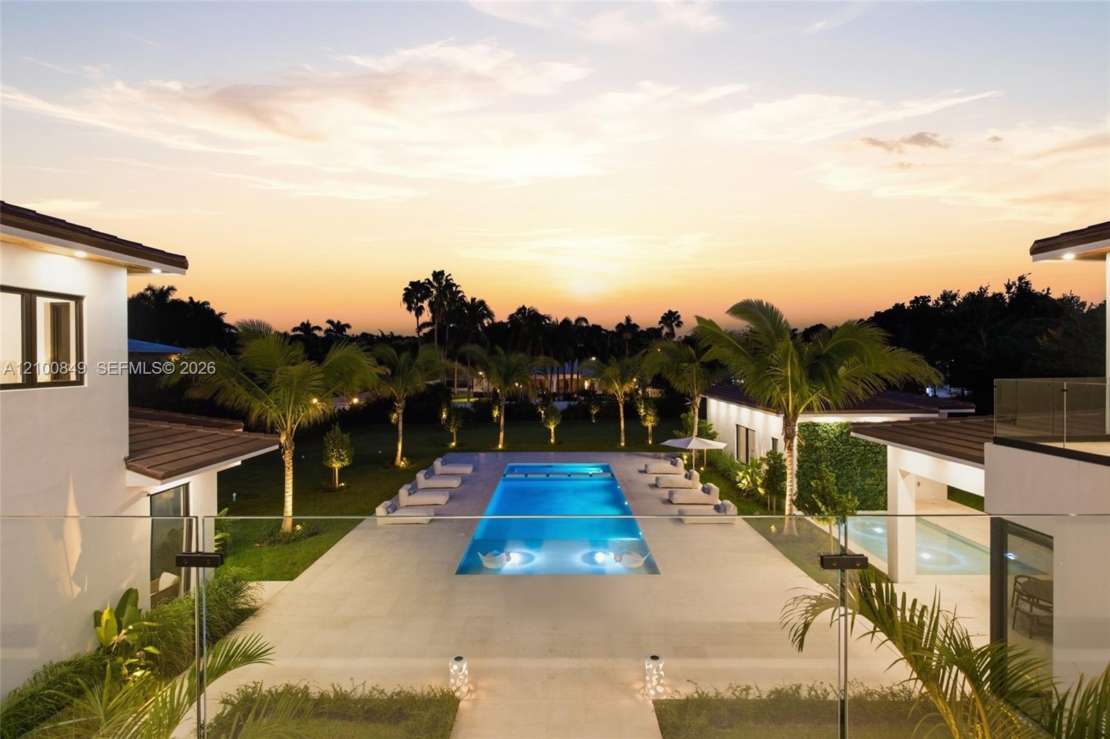 Rectangular swimming pool with integrated sun shelf, expansive stone-paved patio, glass panel perimeter railing, and mature palm landscaping