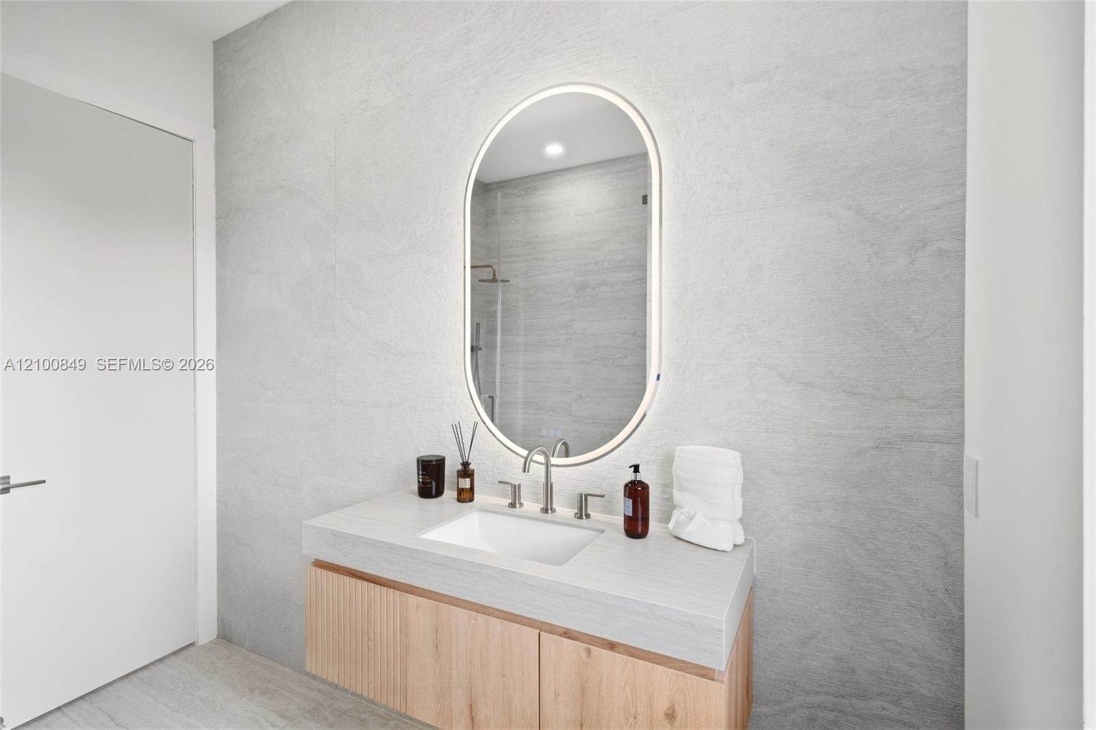 Floating vanity with wood-finish cabinetry, integrated countertop with an undermount sink, wall-mounted faucet, and oval backlit mirror set against te