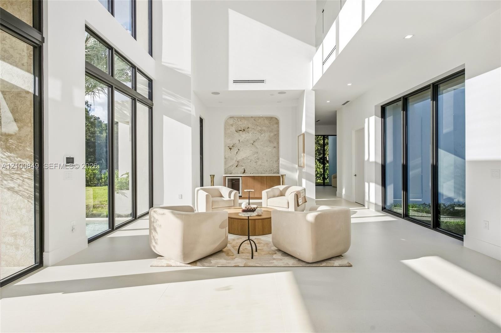 Double-height ceilings, floor-to-ceiling windows with black frames, built-in dry bar with stone backsplash, and smooth-finish flooring