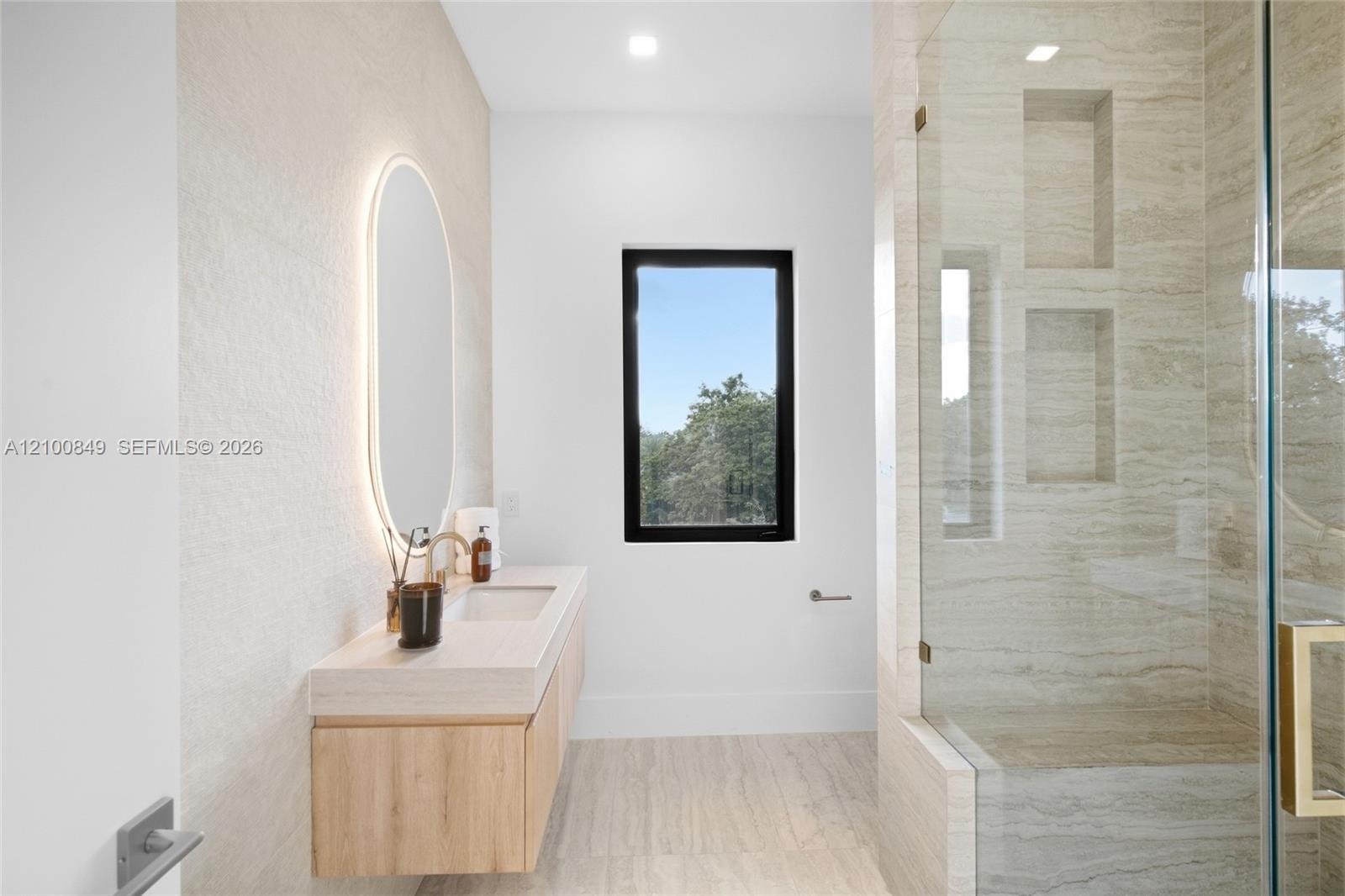 Floating wood-finish vanity with an integrated sink, illuminated oval wall mirror, walk-in shower with stone-tile surround, built-in shower niches, an