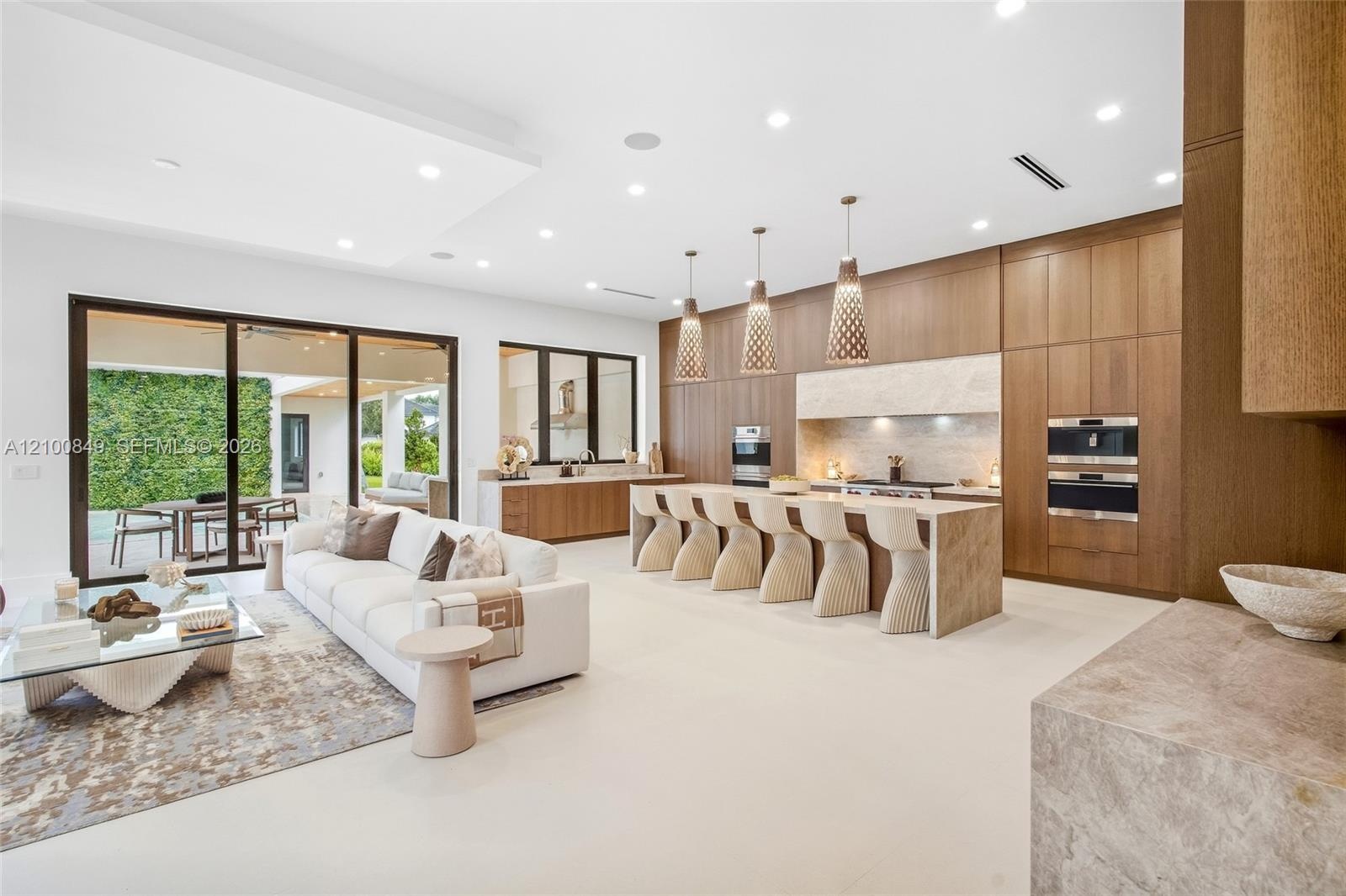 Open-concept living space featuring wood-finish cabinetry, a large waterfall-edge kitchen island, recessed ceiling lighting, and expansive multi-panel