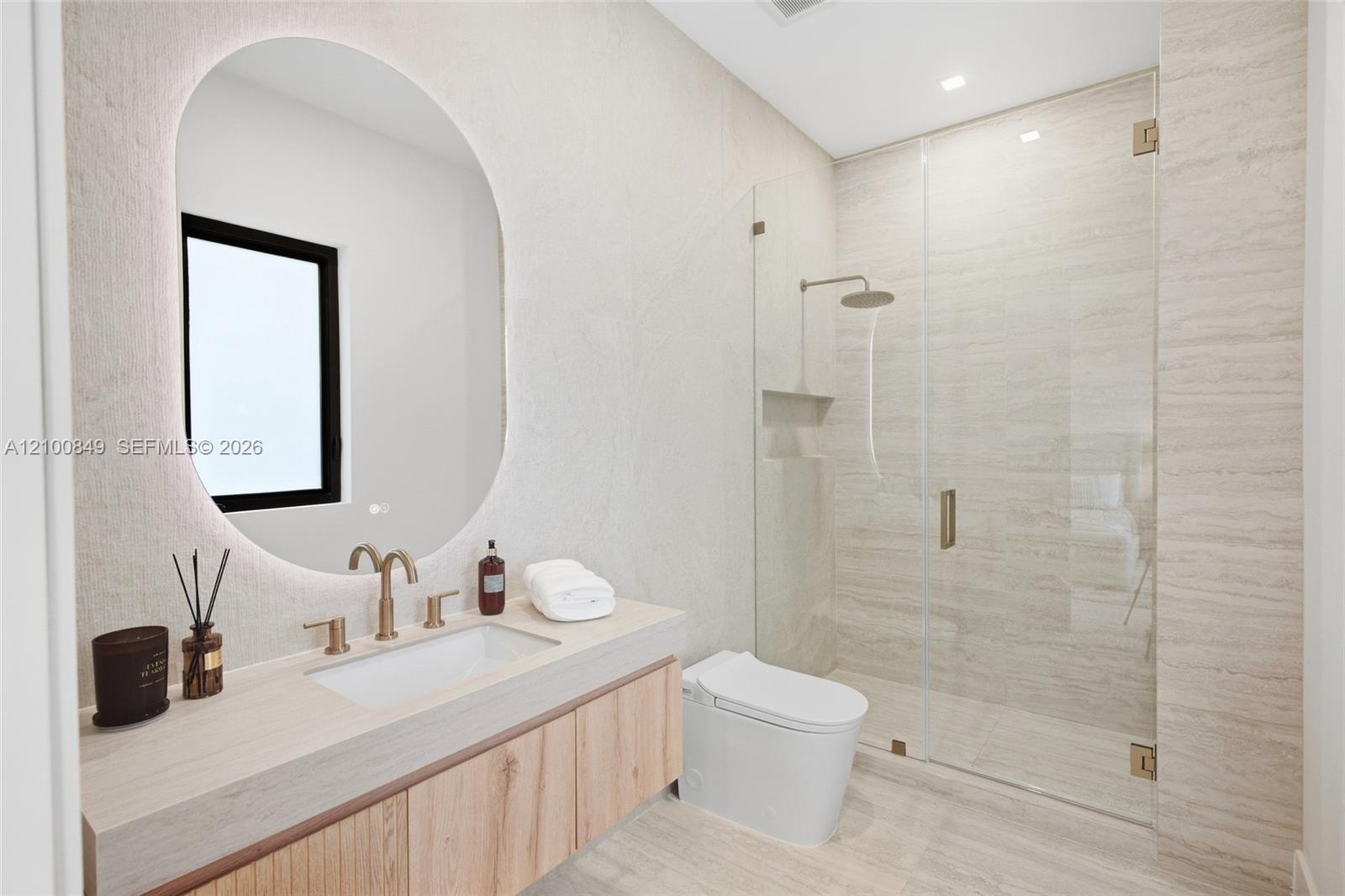 Floating wood-finish vanity with an integrated basin, backlit oval mirror, wall-to-wall stone-finish tiling, and a frameless glass-enclosed shower fea