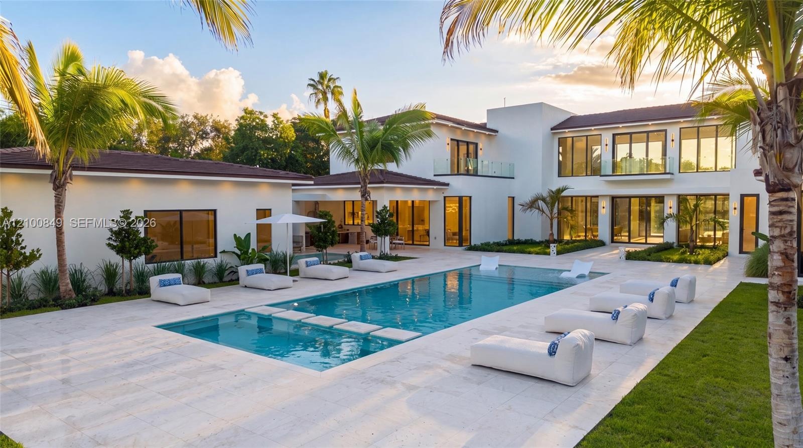 Custom pool with built-in stepping stones, expansive stone-paved patio, two-story white stucco exterior, multiple glass balcony railings, and lush per