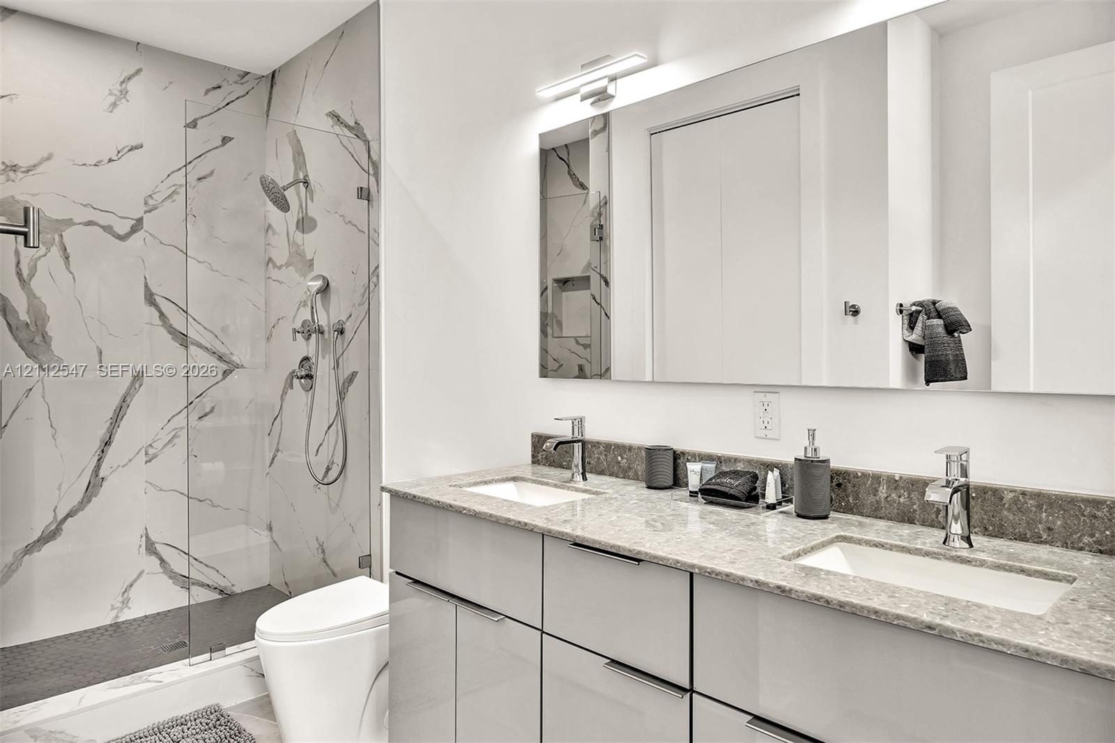 Bathroom featuring a dual-sink vanity with stone countertop, walk-in shower with floor-to-ceiling tile surround, glass partition, and wall-mounted lig