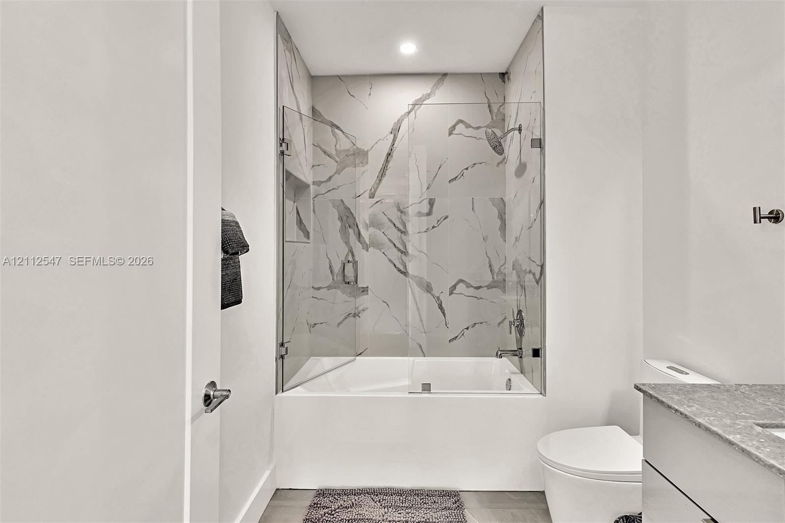 Full-height shower surround with veined tile, frameless glass enclosure, recessed shower niche, deep soaking tub, and stone-finish vanity countertop