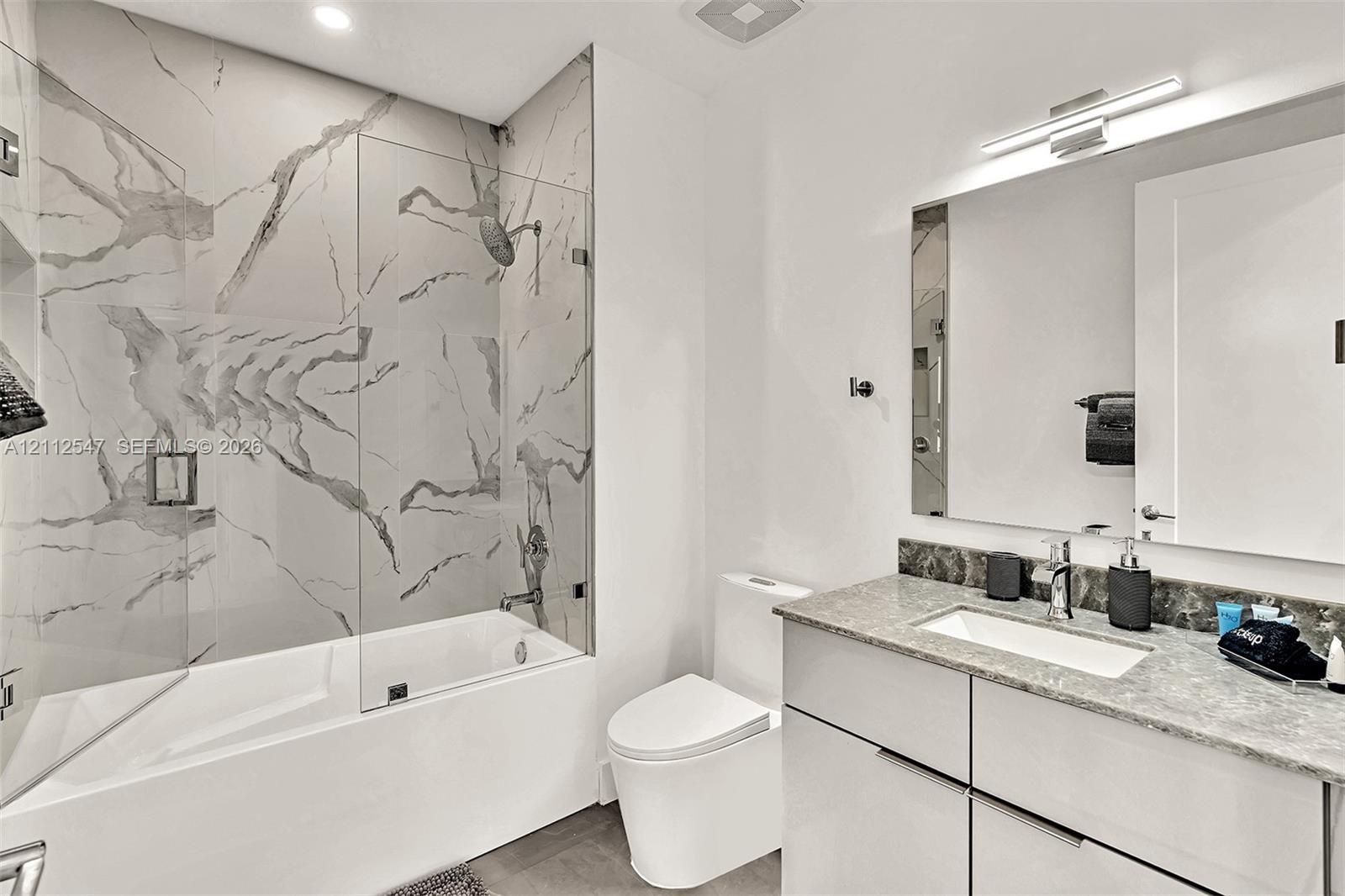 Bathroom featuring a tub-shower combination with floor-to-ceiling large-format tile surround, a glass shower panel, a floating vanity with a stone cou
