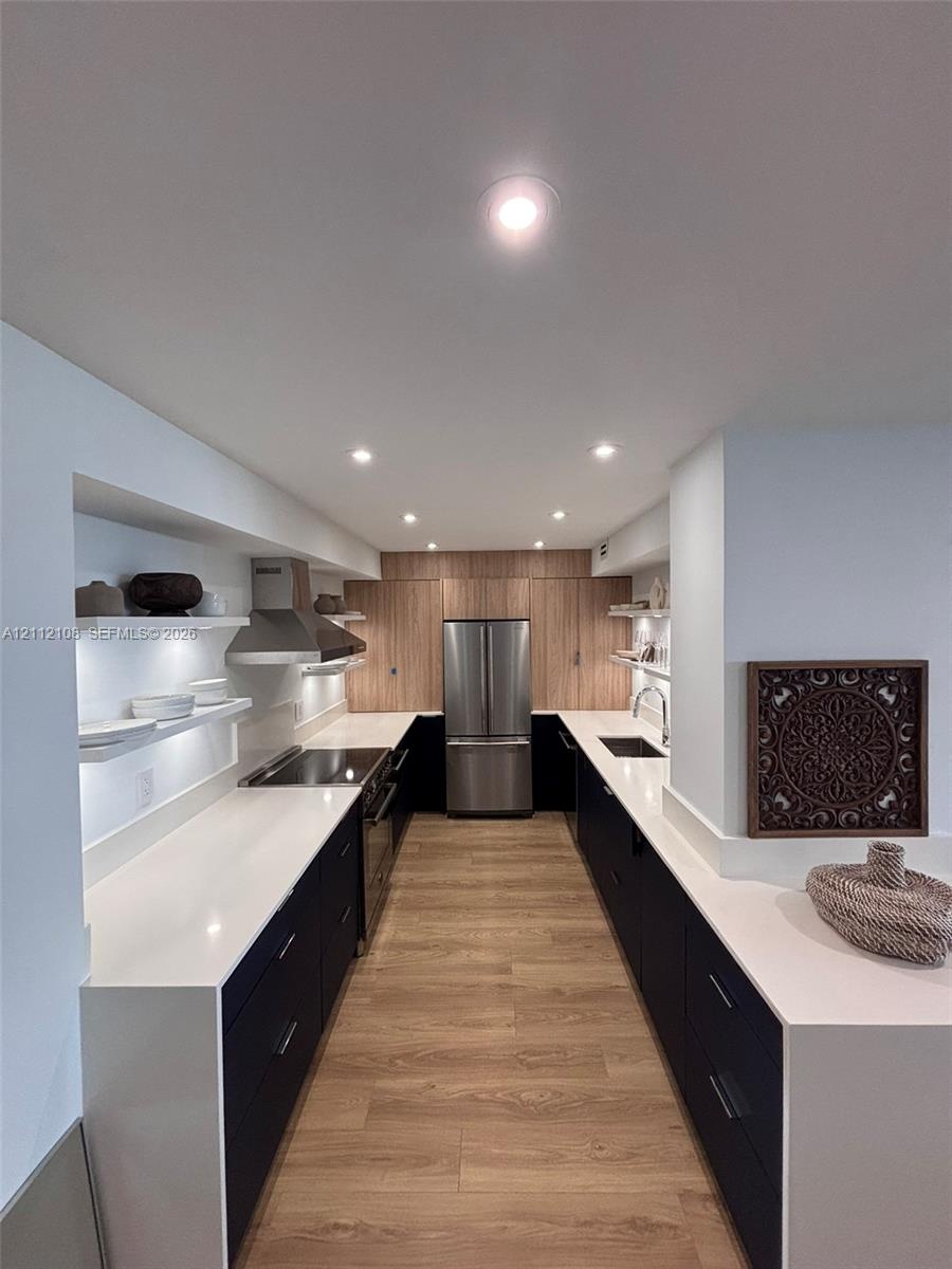 Kitchen featuring dark flat-panel cabinetry, light-toned stone countertops, wood-finish flooring, stainless steel appliances, and integrated recessed 