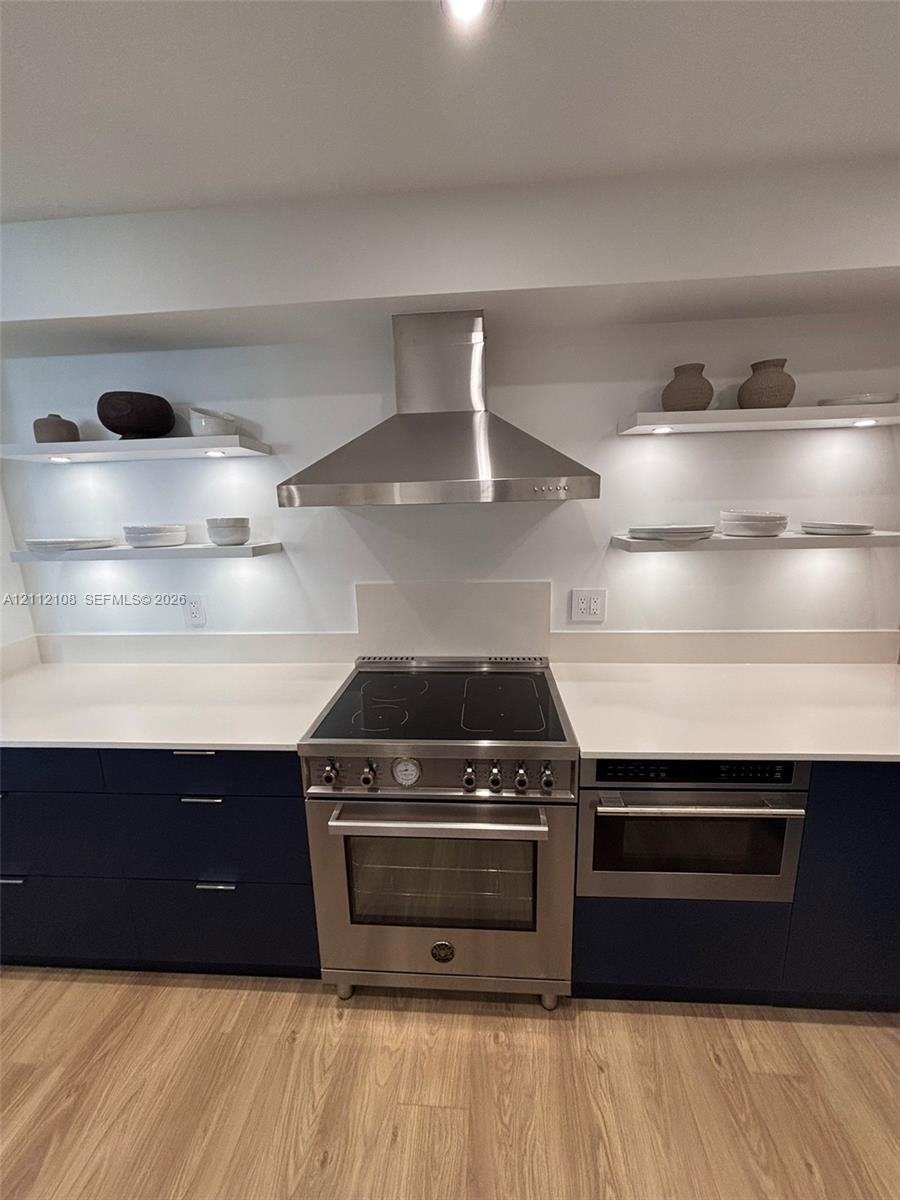 Kitchen featuring dark flat-panel cabinetry, quartz countertops, a stainless steel range with a matching chimney-style hood, and integrated under-shel